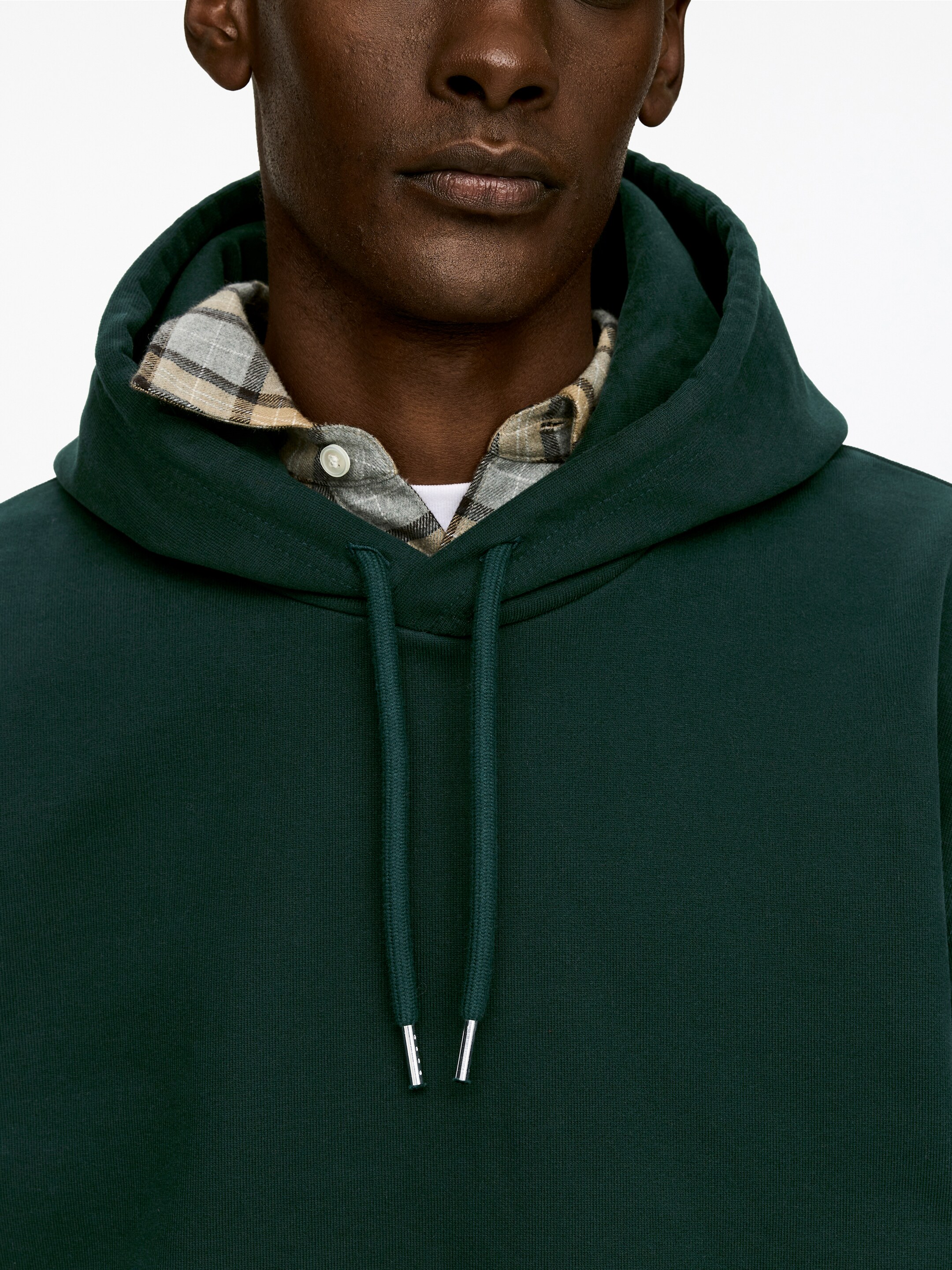 Ver imagem maior: Uma camisola com capuz verde escuro está a ser apresentada de um ângulo frontal, em close-up. Apresenta um capuz visível e cordões verdes escuros com terminais prateados, e está sobreposta a uma camisa de xadrez com a gola visível.