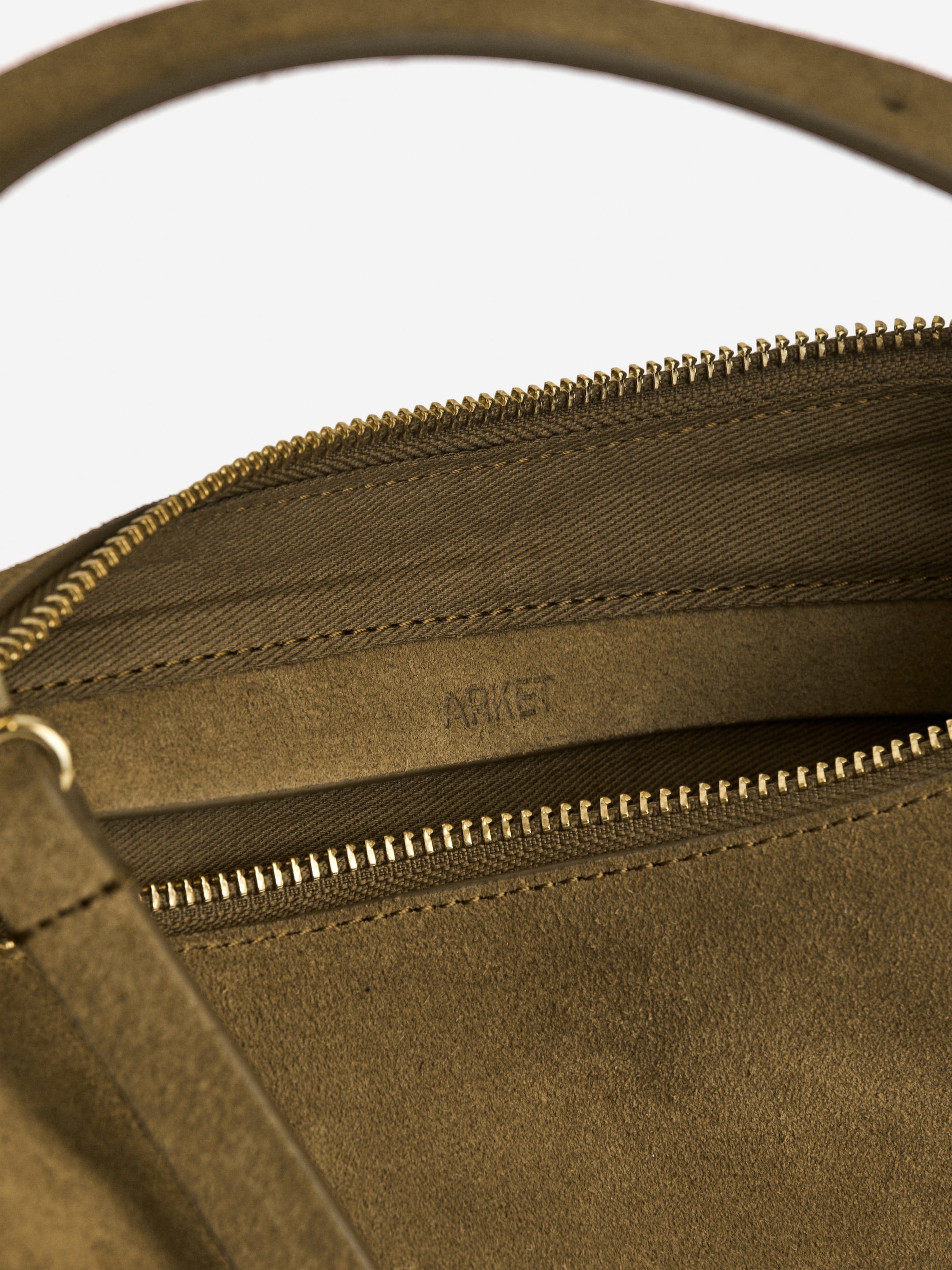 Grösseres Bild ansehen: Eine olivgrüne Ledertasche wird aus einer Nahaufnahme von oben gezeigt, wobei ihr offenes Hauptfach mit einem goldfarbenen Reißverschluss zu sehen ist. Ein gebogener Tragegriff ist teilweise sichtbar, und im Inneren werden eine Innentasche mit einem passenden goldfarbenen Reißverschluss und das eingeprägte 'ARKET'-Branding sichtbar.