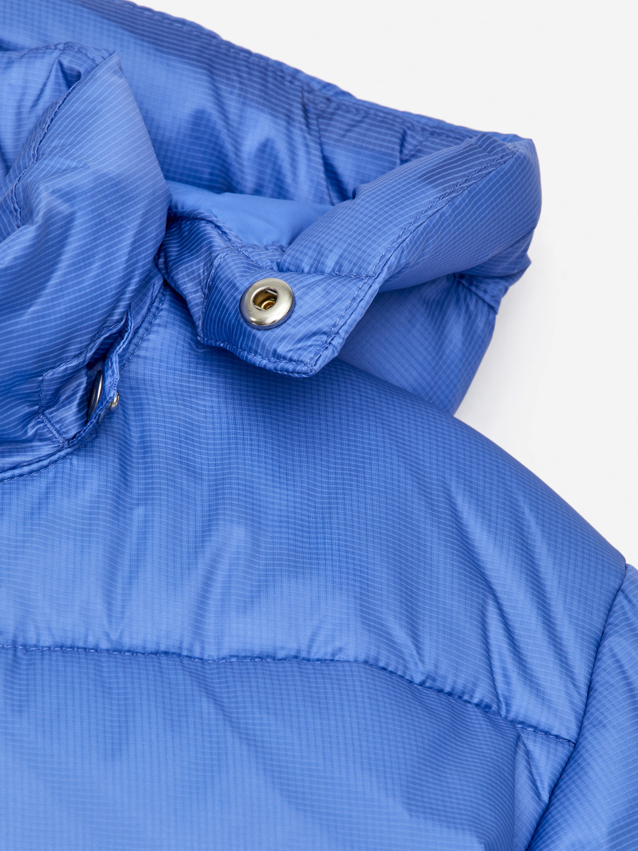Agrandir l'image: Une veste matelassée bleue est présentée, mettant en valeur sa texture matelassée et un motif en grille subtil sur le tissu. Une capuche amovible est fixée par un bouton-pression en métal visible.