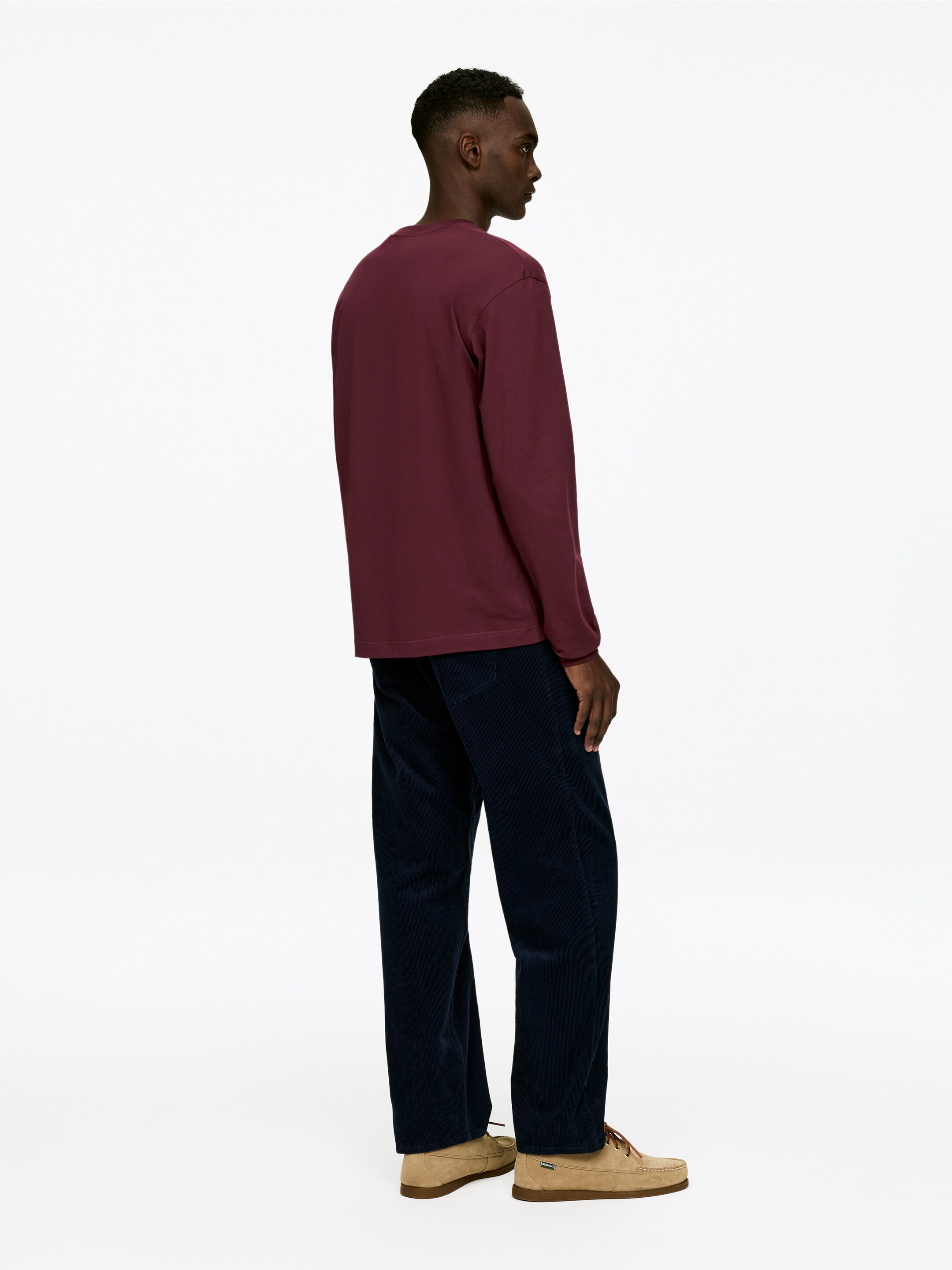 Agrandir l'image: Un t-shirt bordeaux à manches longues et col rond est présenté en vue arrière trois-quarts, avec une coupe décontractée, des poignets simples et un ourlet droit qui tombe sur un pantalon en velours côtelé bleu foncé.