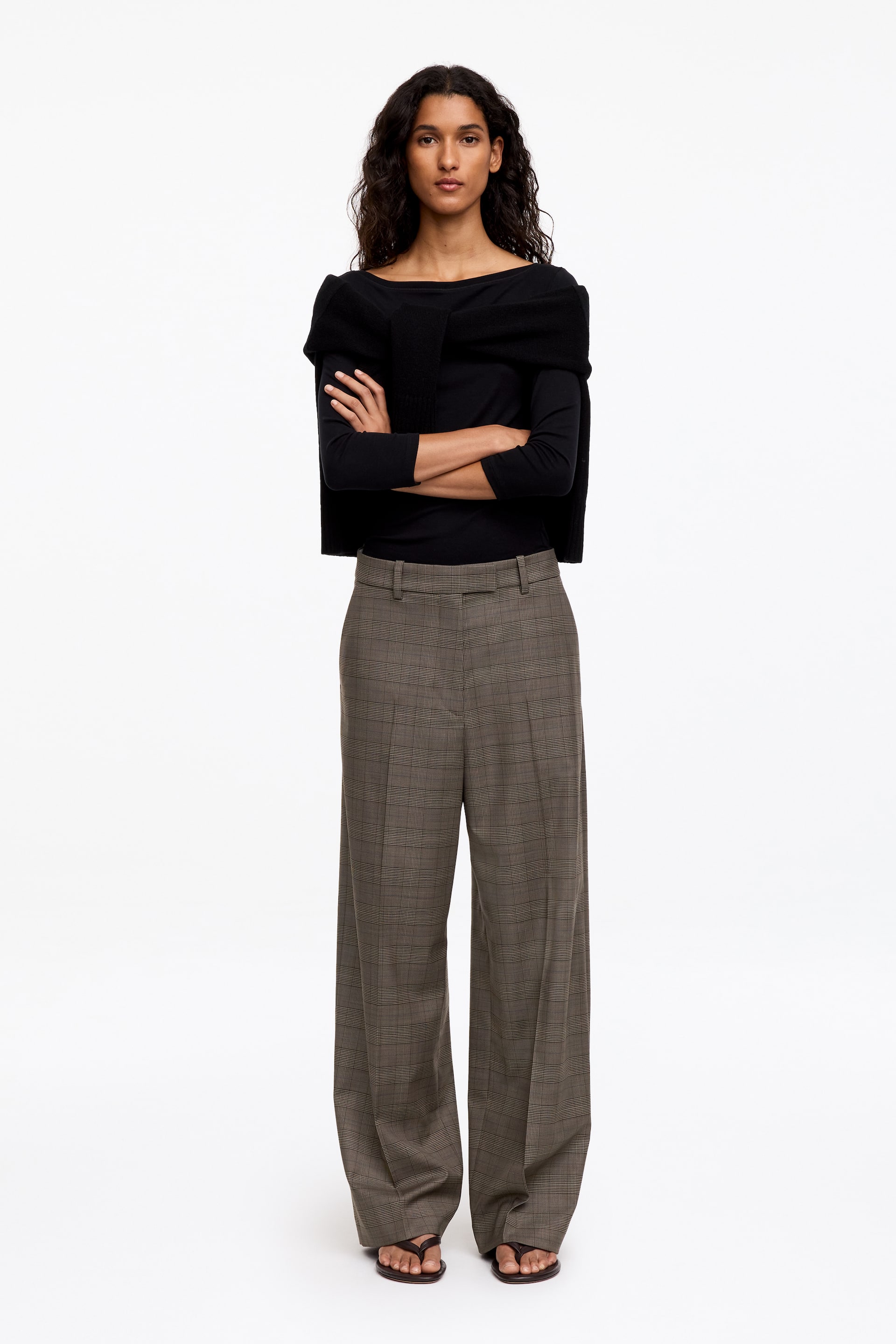 Grotere afbeelding bekijken: De model staat met gekruiste armen en draagt een op maat gemaakte geruite broek met een relaxte, rechte pijp en strakke persvouwen aan de voorkant. De broek heeft een nette tailleband met riemlussen en is gecombineerd met een zwarte top met lange mouwen, een zwarte gebreide trui over de schouders en donkere platte sandalen.