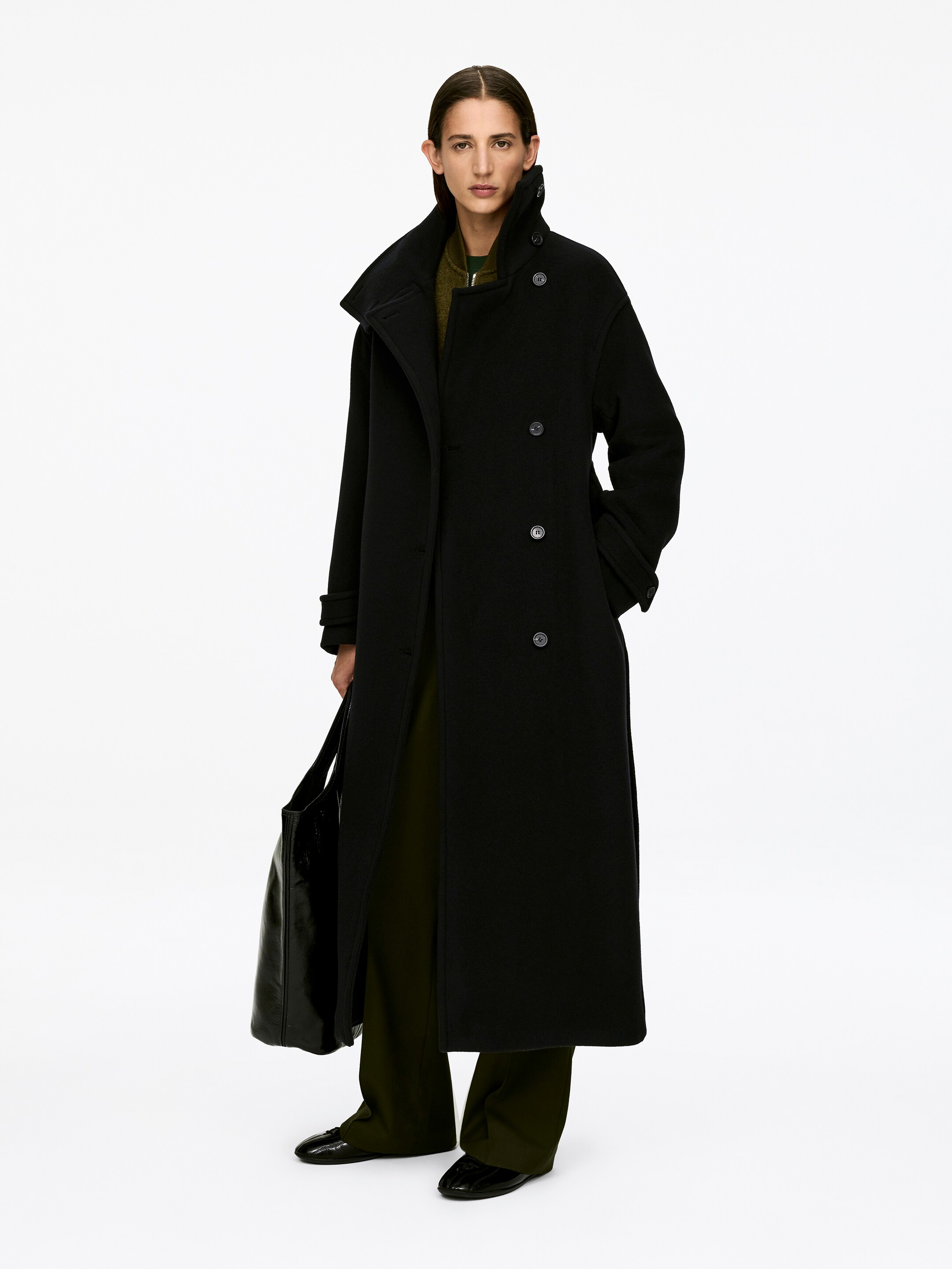Se større billede: En sort, oversized maxi-frakke i uldblanding, med en stående krave, en dobbelt-breasted front med to søjler af knapper, og ærmefaner med knaplukninger, bæres af modellen over en grøn top og bukser. Modellens venstre hånd hviler i en sidelomme, og en sort taske bæres i højre hånd.