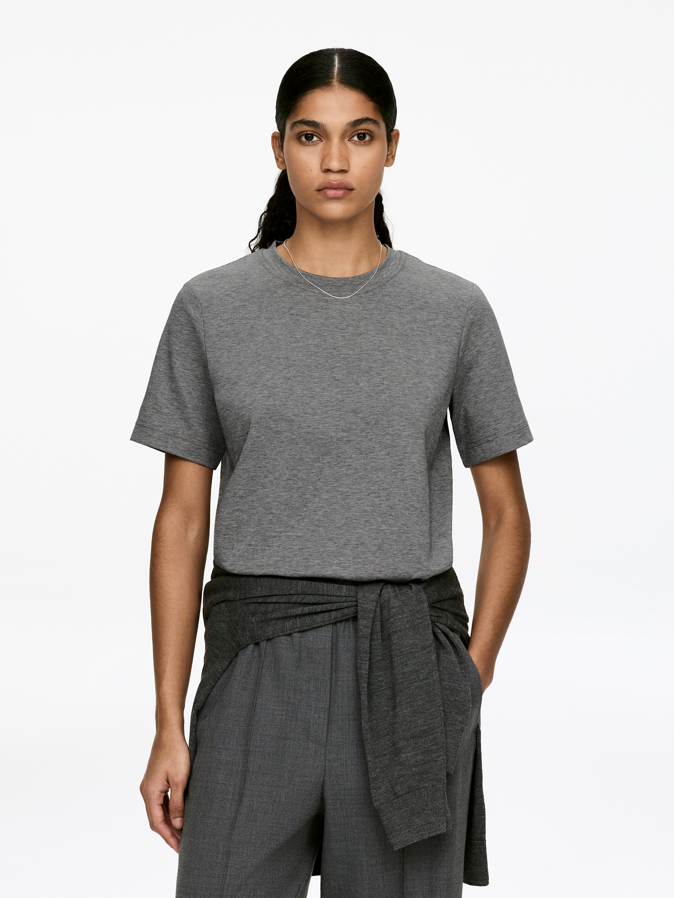 View larger image: A gray cotton jersey T-shirt, featuring a classic crew neck and short, softly tailored sleeves, is being worn by the model. A thin silver necklace is resting on the T-shirt, and a dark gray garment is tied around the model's waist.