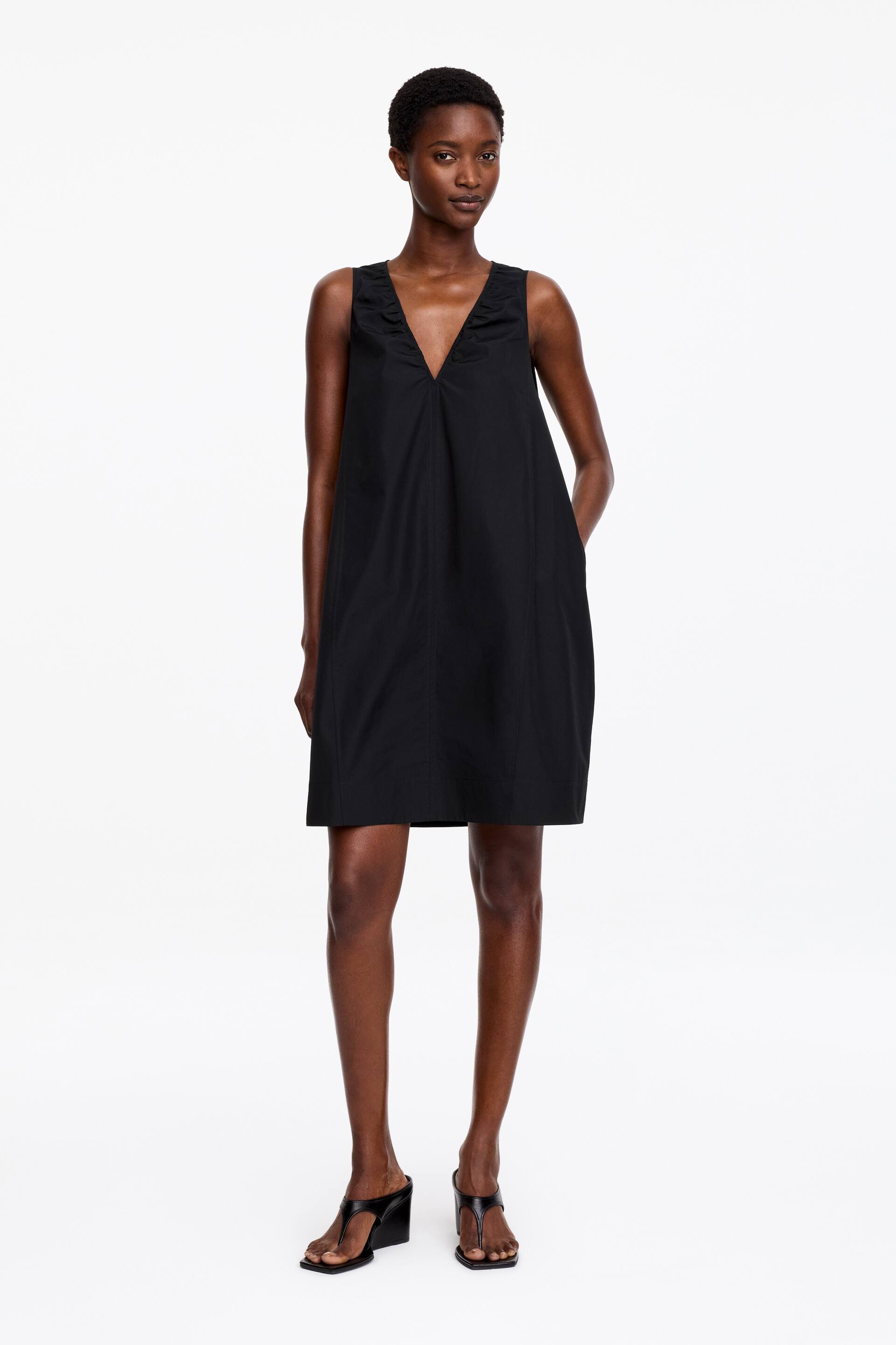 Größeres Bild ansehen: Schwarzes Kleid mit tiefem V-Ausschnitt und gerafften Details sowie ärmellosen Armlöchern. Es präsentiert eine entspannte A-Linien-Silhouette mit vertikalen Nähten, die die Vorderseite definieren, und ist mit sichtbaren Seitentaschen gestaltet, endet oberhalb des Knies.