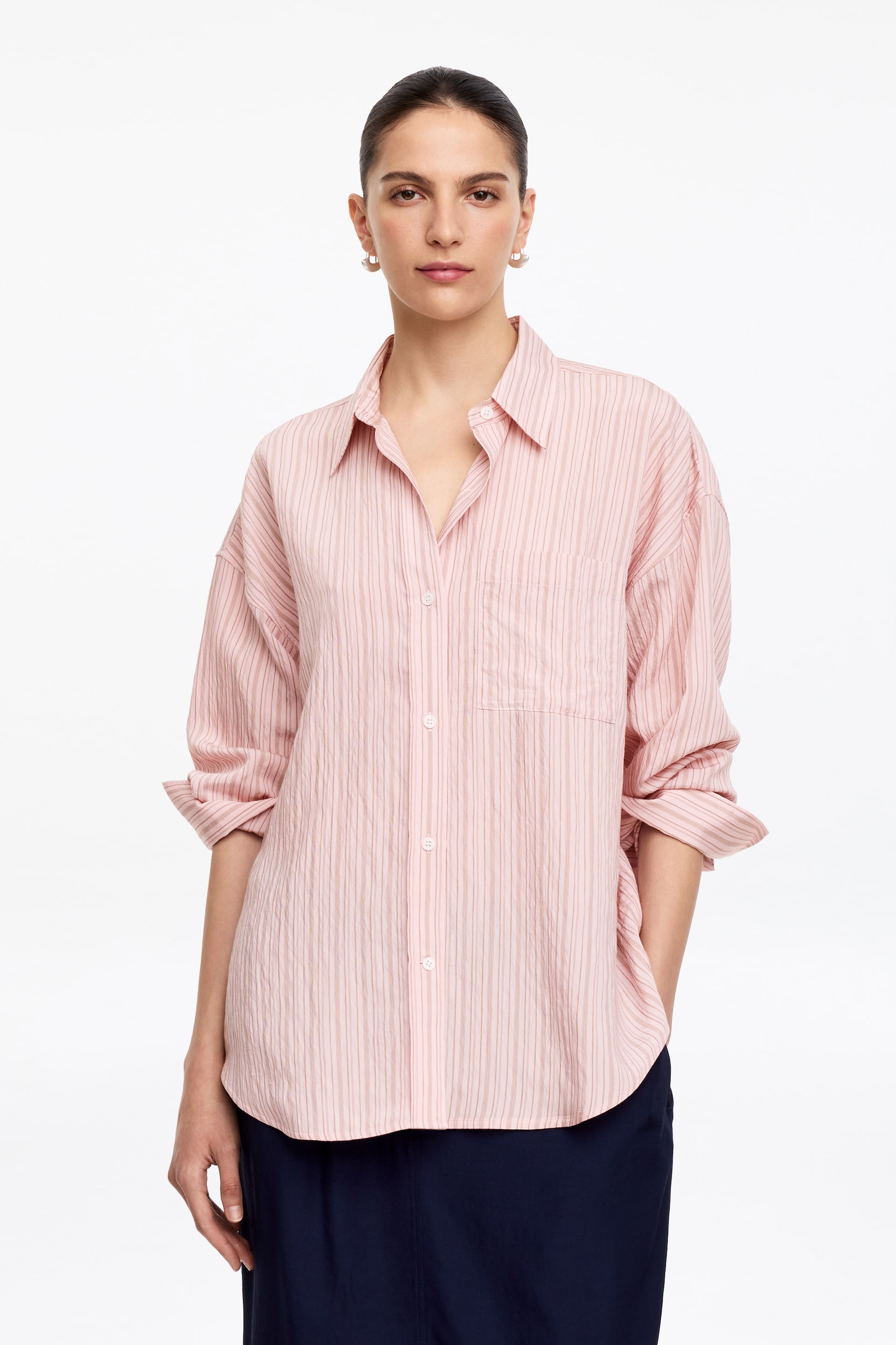 Visualizza immagine più grande: Una camicia a righe rosa con bottoni è presentata, rivolta in avanti. Presenta un colletto a punta, una chiusura frontale con bottoni bianchi e una singola tasca applicata sul petto sinistro, mostrando una texture stropicciata e righe verticali, con le maniche lunghe arrotolate.
