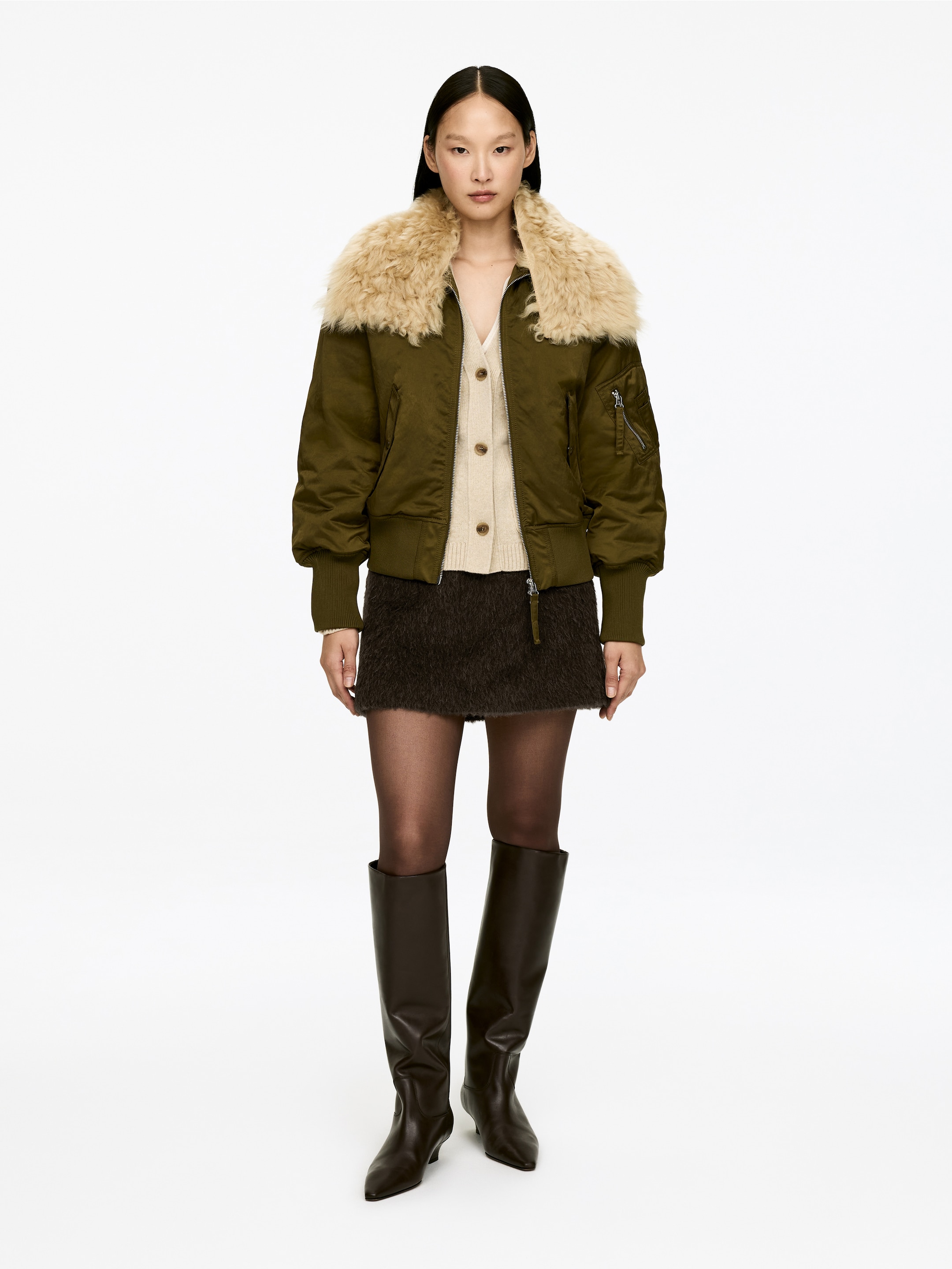 Agrandir l'image: Une veste bomber vert olive est portée, face avant, avec un grand col en fourrure beige clair et duveteux. Elle présente des poignets et un ourlet côtelés, ainsi qu'une poche zippée sur la manche gauche, tout en étant superposée à un cardigan beige clair.