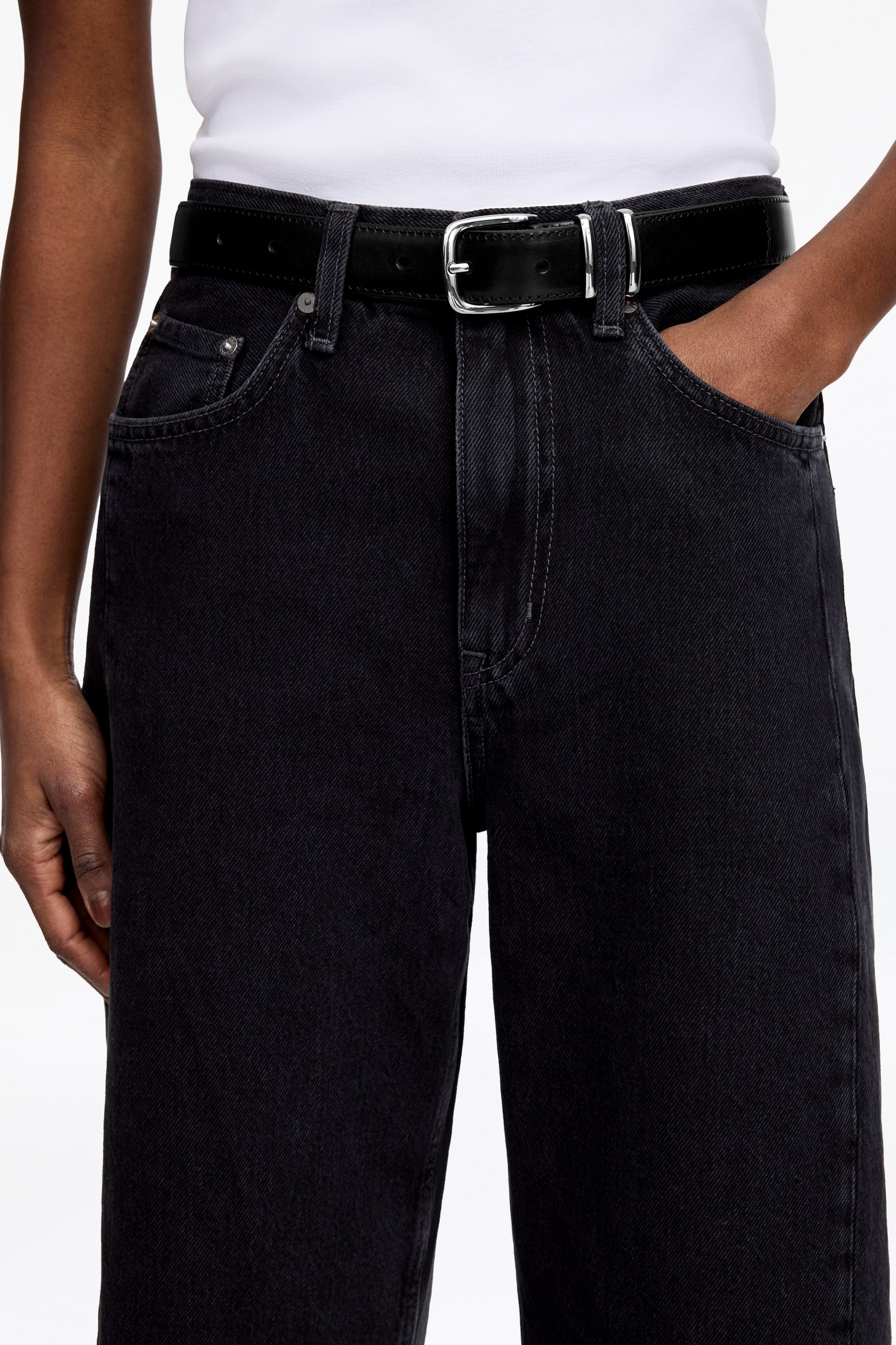 Größeres Bild ansehen: Schwarze Denim-Jeans werden präsentiert, mit hoher Taille, sichtbaren Fronttaschen mit Metallnieten und kontrastierenden Nähten. Ein schwarzer Ledergürtel mit einer silberfarbenen rechteckigen Schnalle ist durch die Gürtelschlaufen gezogen und fixiert die Jeans in der Taille.