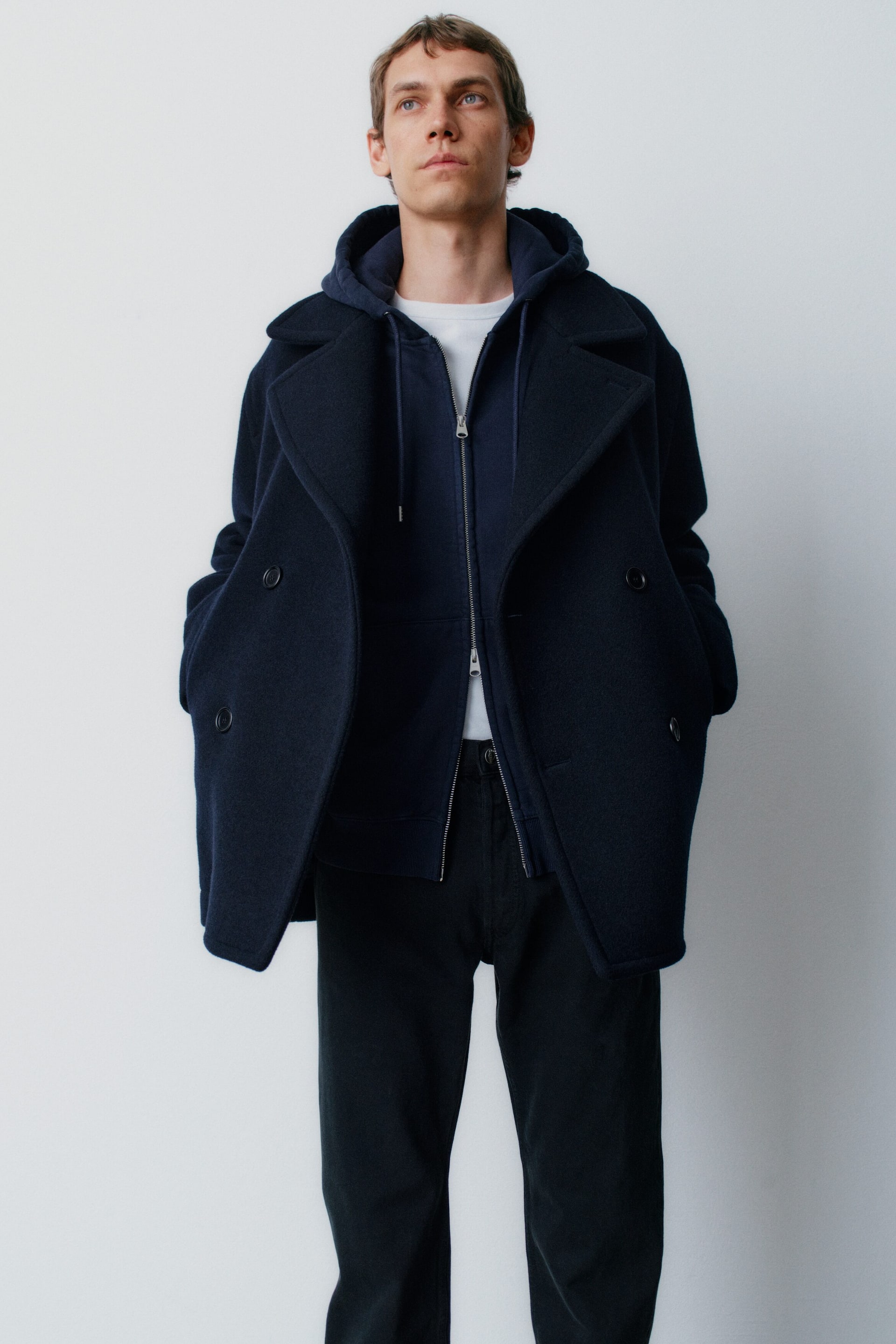 Se større billede: Viser et frontalt billede er modellen iført en mørk marineblå pea coat i uldblanding med brede revers, båret over en marineblå French terry-hoodie med tovejslynlås og en hvid tætsiddende rib T-shirt. Looket fuldendes med sorte, lige benede bukser i uld og bomuld.