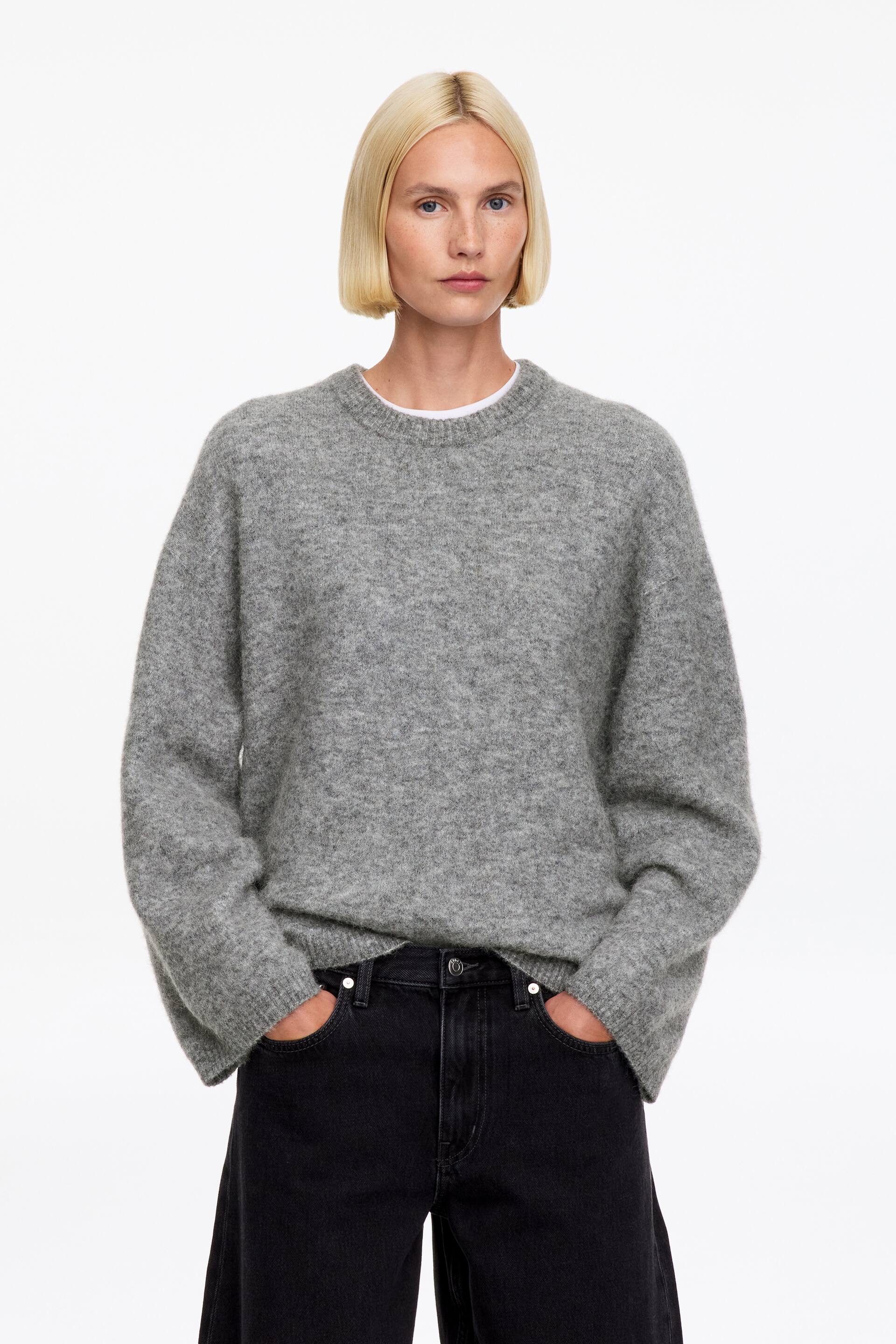 Visa större bild: En grå, strukturerad stickad tröja med rund hals bärs av modellen över en lagerlagd vit T-shirt. Tröjan med avslappnad passform har långa, vida ärmar och är stylad med svarta denimjeans.