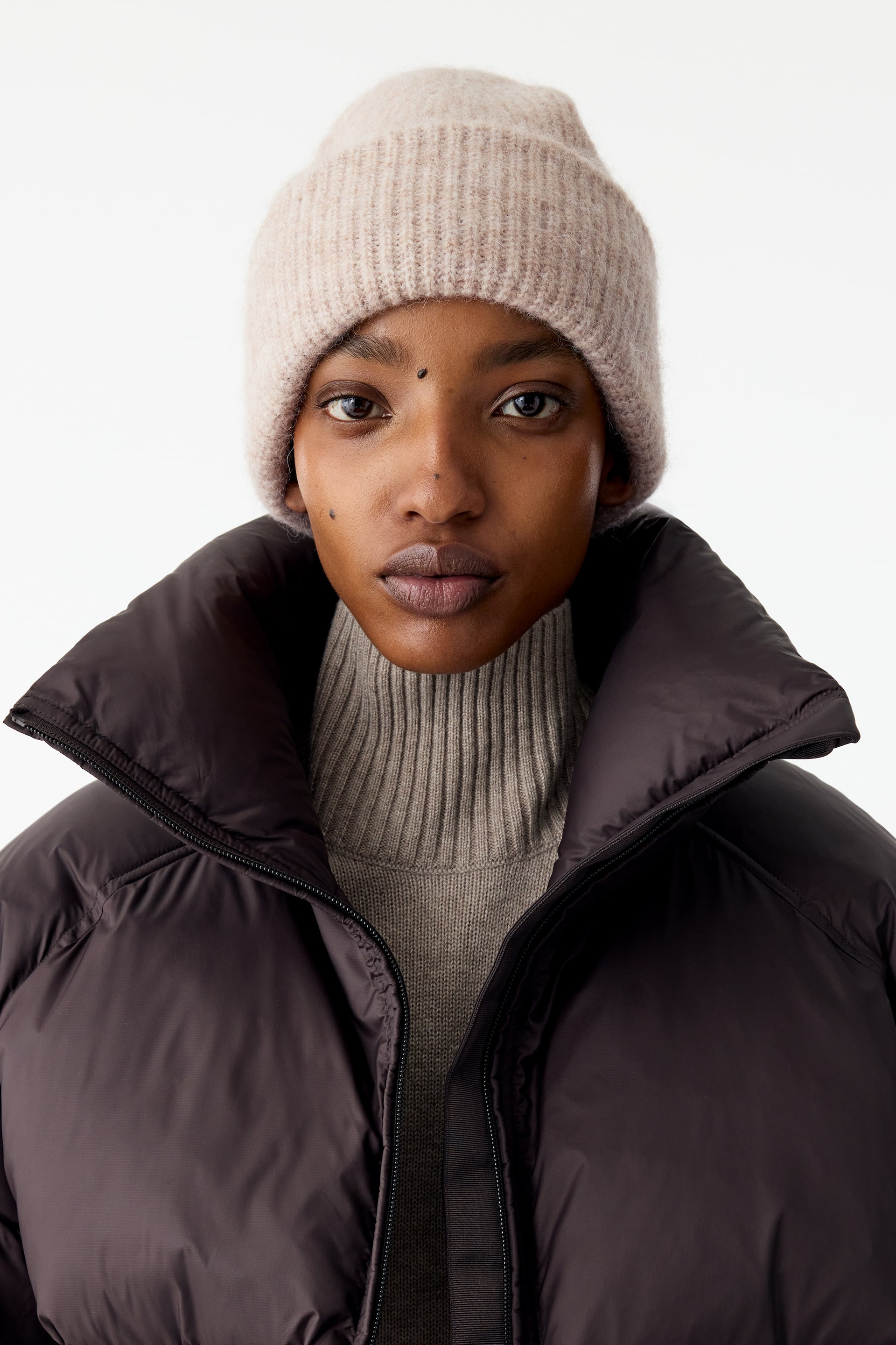 Größeres Bild anzeigen: Eine dunkelbraune Pufferjacke mit hohem Stehkragen wird vom Model offen getragen und über einem taupefarbenen Rollkragenpullover aus Rippstrick kombiniert. Auf dem Kopf trägt das Model zudem eine hellbeige Rippstrick-Mütze, die das winterliche Layering abrundet.