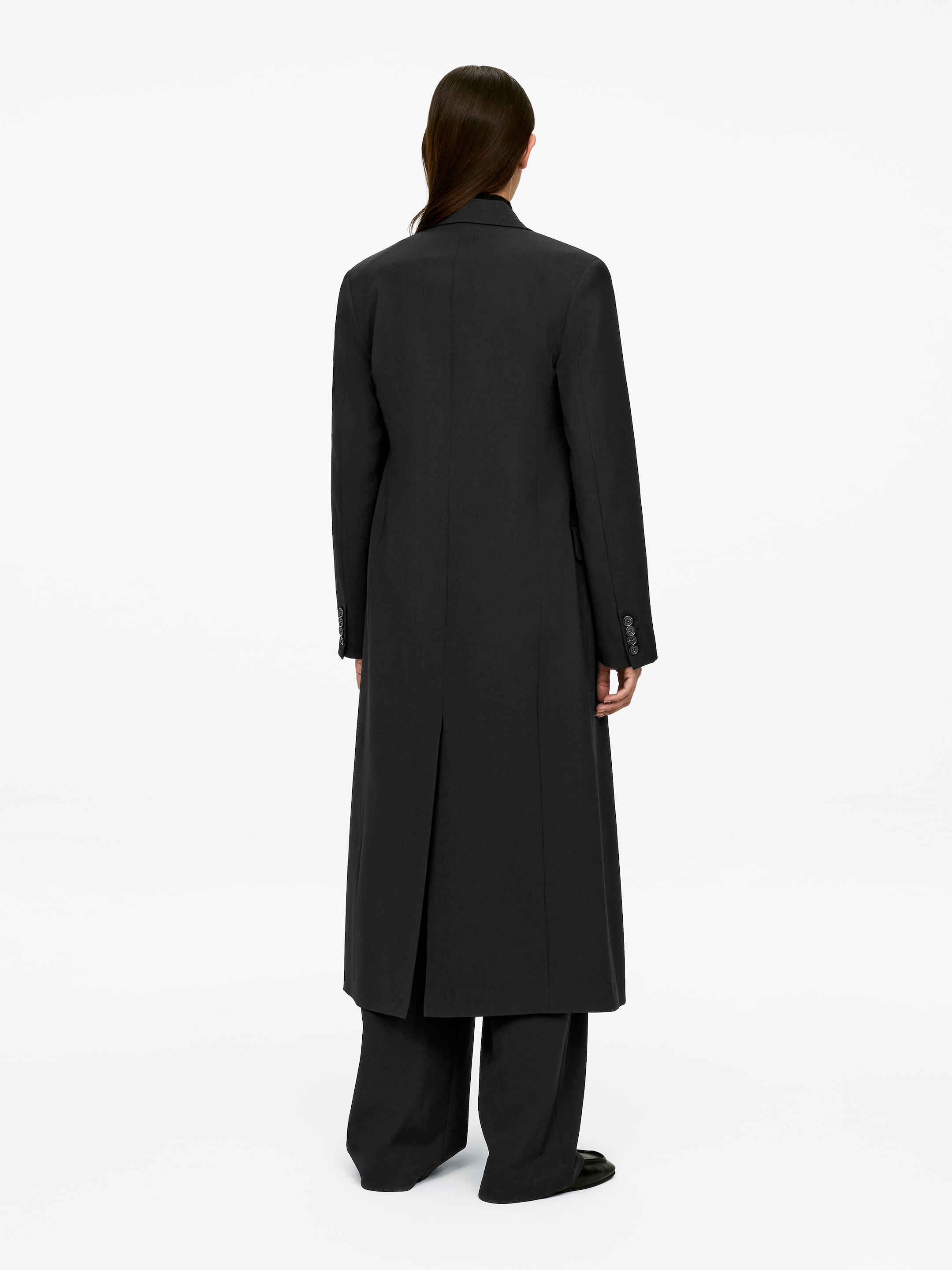 Visa större bild: En lång svart kappa med strukturerade axlar, knäppta manschetter och en mitt-bak-sprund bärs av modellen, som står med ryggen mot betraktaren. Modellen har också på sig svarta byxor med breda ben och svarta skor.