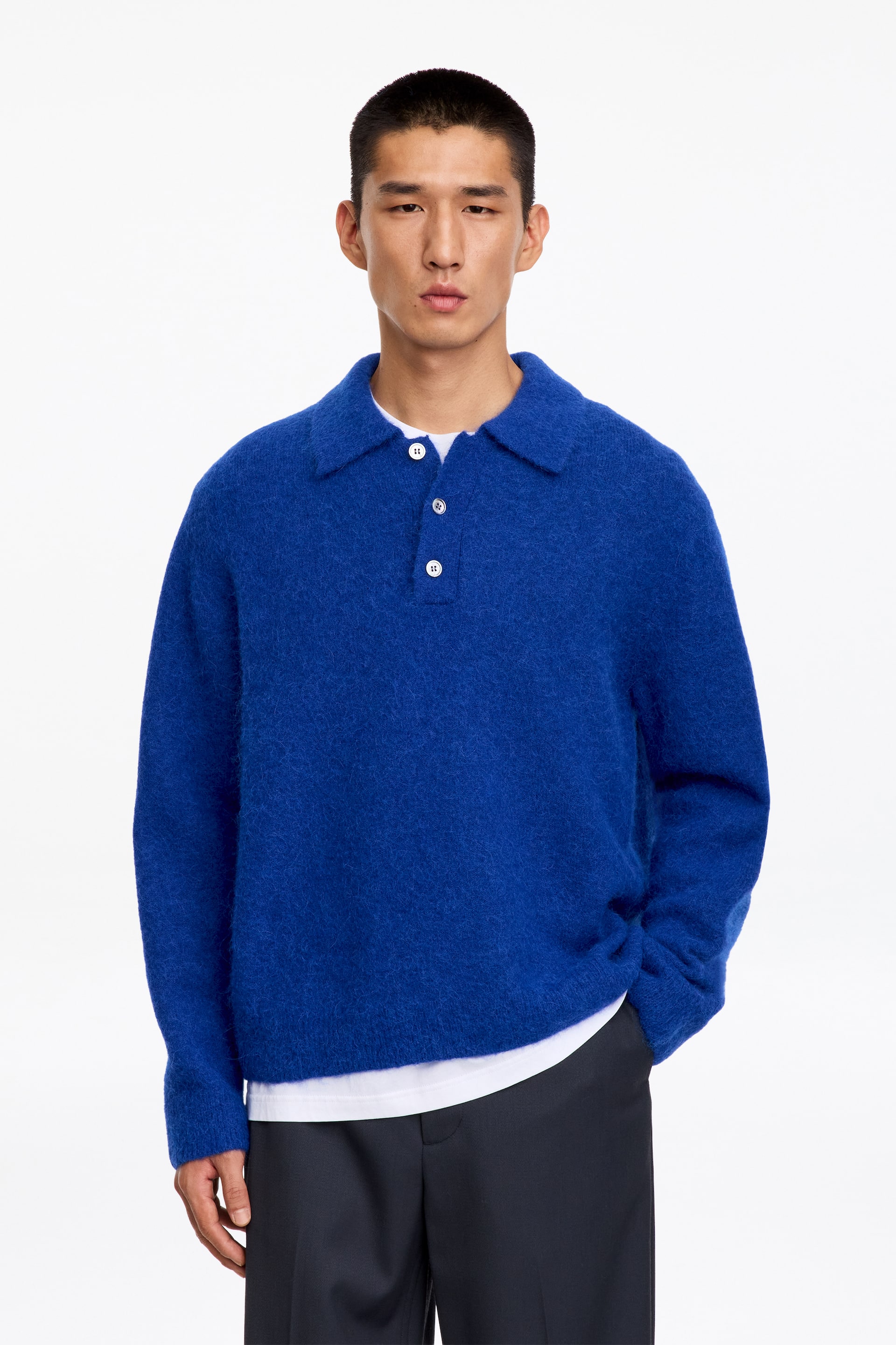 Größeres Bild ansehen: Ein leuchtend blauer, flauschiger Strick-Polopullover mit Kragen und Drei-Knopf-Leiste wird vom Model getragen. Der Pullover wird über einem weißen T-Shirt getragen und mit dunklen Hosen kombiniert.