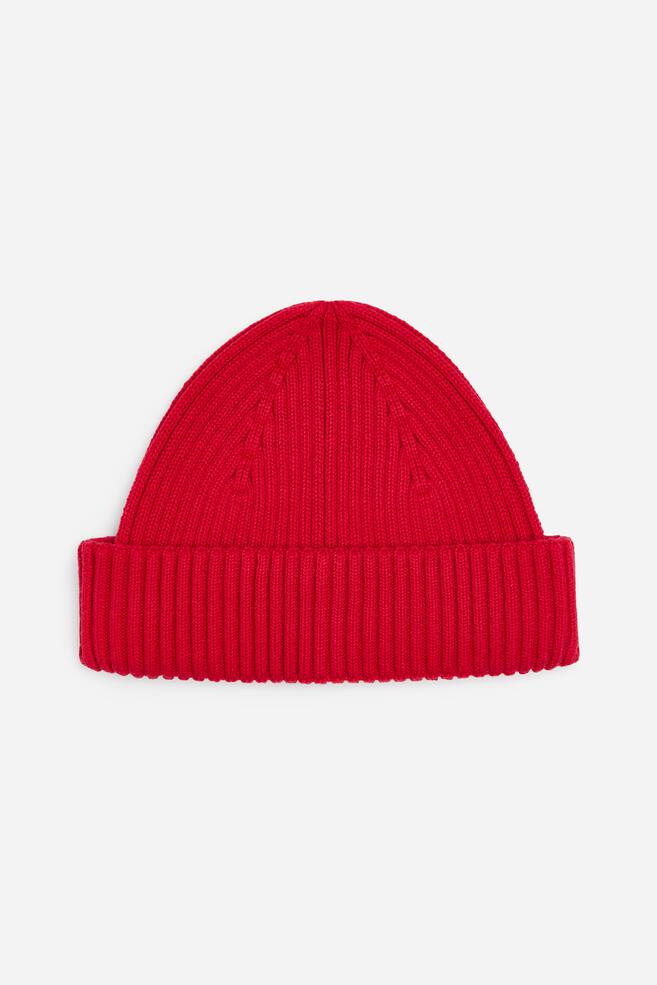 A red rib-knitted beanie is shown facing front, featuring a vertically ribbed texture and a wide, folded brim. - 1