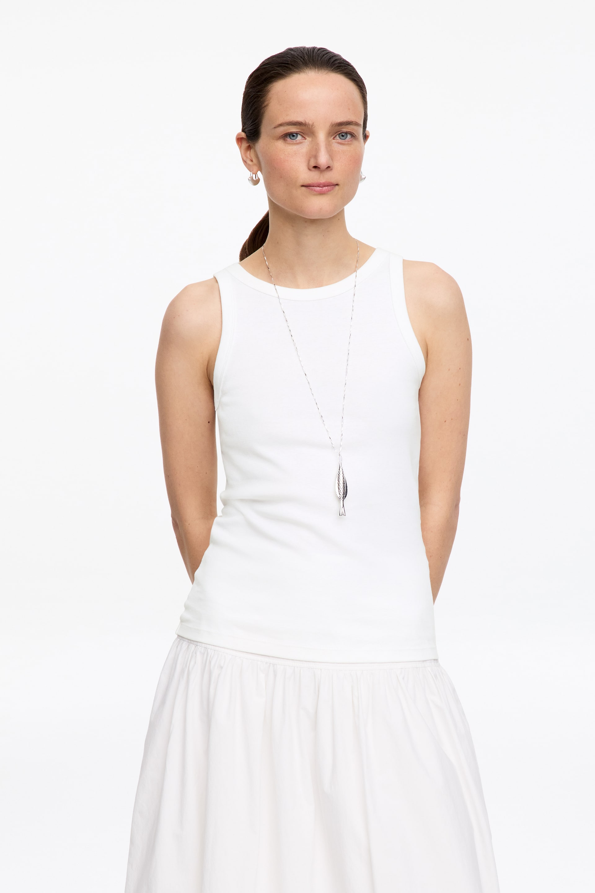 Visa större bild: En vit ribbad tanktopp med rund hals och ärmlös design bärs tillsammans med en vit rynkad kjol. Ett långt silverkedjehalsband med ett fiskhänge är lager på lager över tanktoppen, och små silverringar syns också.
