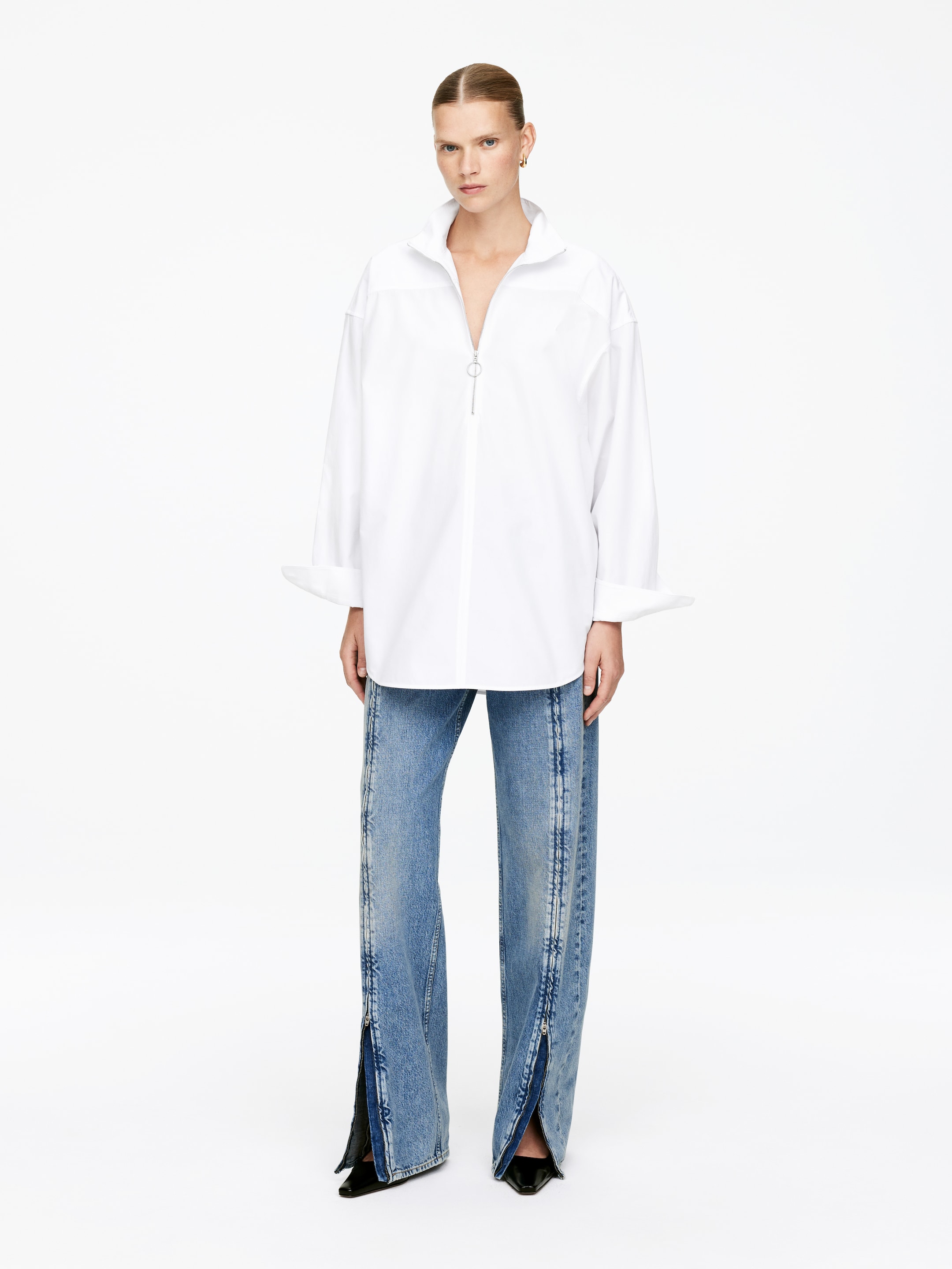 Visa större bild: En överdimensionerad vit skjorta med ståkrage, en halv dragkedja framtill med ett runt drag och breda uppvikta manschetter bärs tillsammans med ljusblå jeans. Jeansen har framträdande vertikala sömmar och fullängds sidodragkedjor som är delvis uppdragna vid fållen.