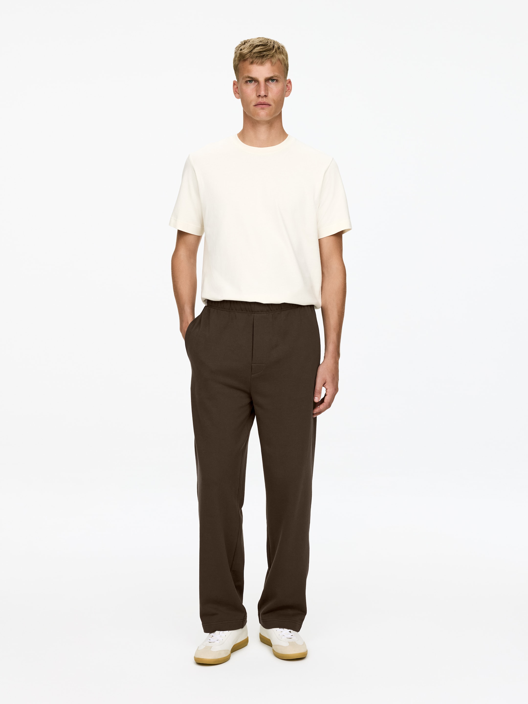 Ingrandisci l'immagine: Pantaloni della tuta marrone scuro, tagliati per una vestibilità regolare, sono indossati dal modello. Presentano una cintura elasticizzata con un laccio visibile e tasche laterali, e il modello sta mettendo una mano in una tasca laterale.