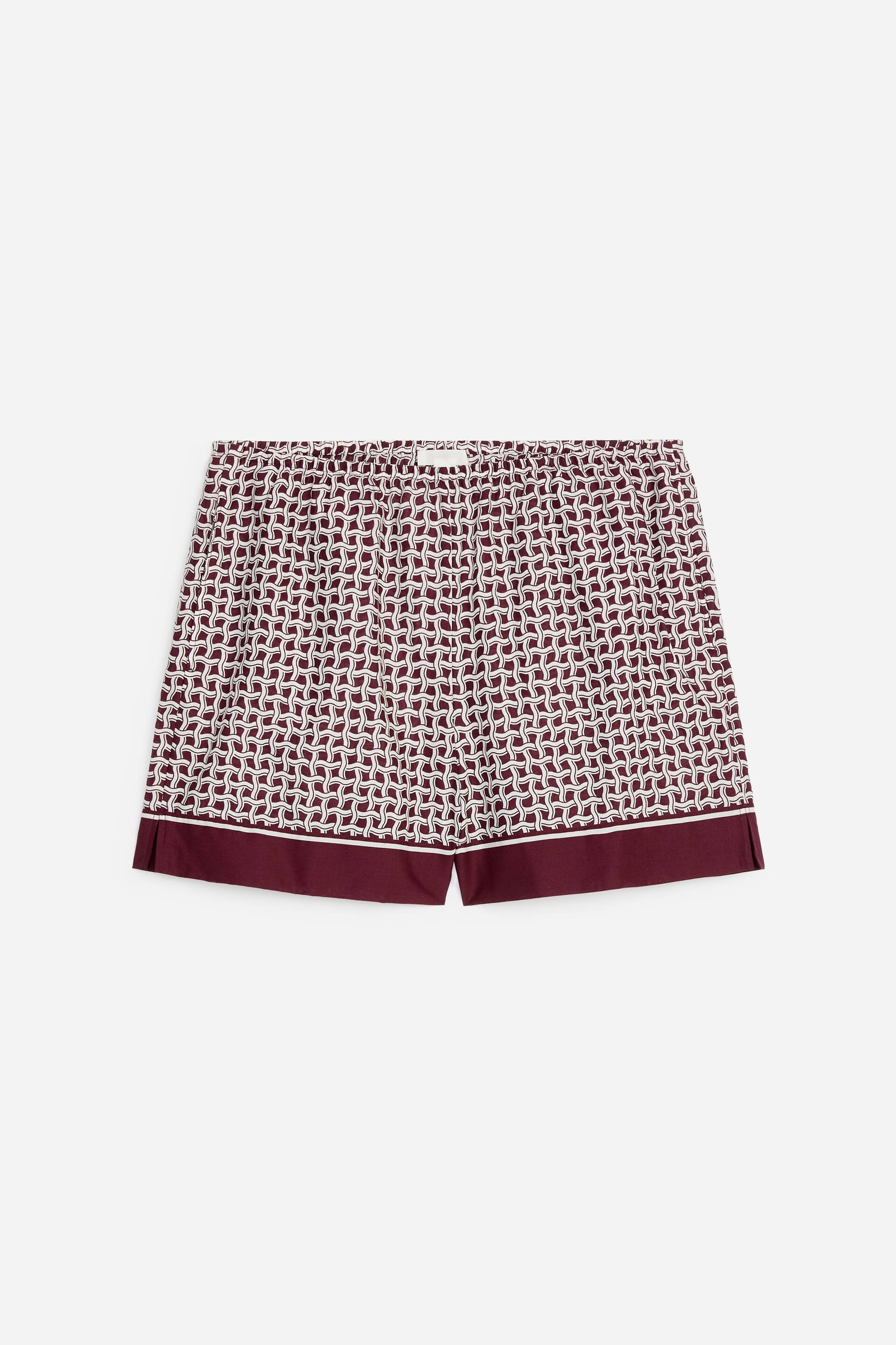 Grösseres Bild ansehen: Ein Paar Shorts zeigt seine Vorderseite mit einem durchgehenden geometrischen Kettengliedmuster in Kastanienbraun und Weiß. Sie sind mit einem elastischen Bund gestaltet und auf eine knielange Silhouette zugeschnitten, mit einem soliden kastanienbraunen Band, einem dünnen weißen Streifen und kleinen seitlichen Schlitzen, die den Saum detaillieren.
