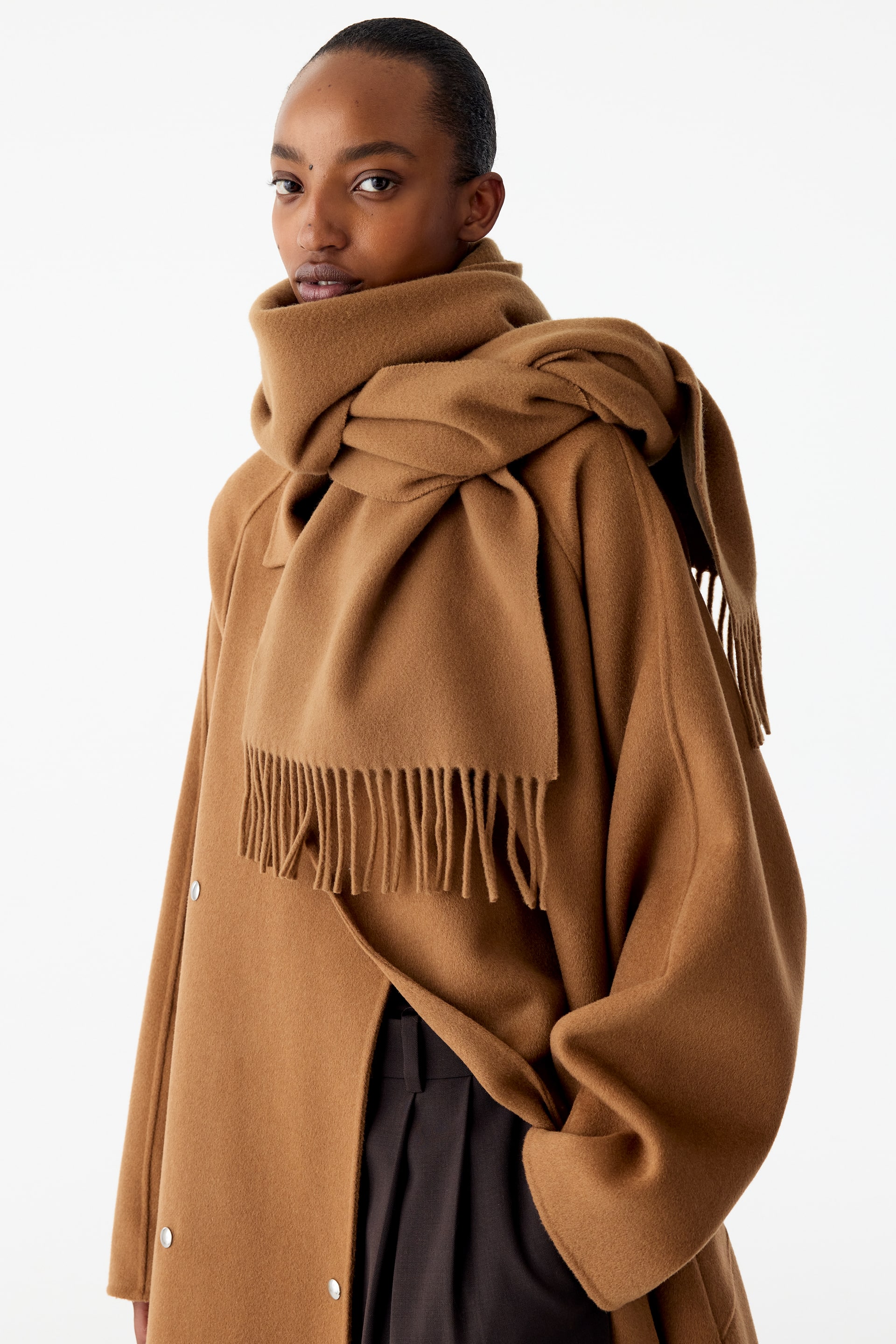 Visualizza immagine più grande: La modella indossa una sciarpa in lana color cammello, avvolta spessamente intorno al collo e adagiata sopra un cappotto coordinato. La sciarpa, tessuta fitto, presenta lunghe frange sottili lungo i bordi più corti, con un’estremità che pende sul davanti e l’altra drappeggiata sulla spalla.