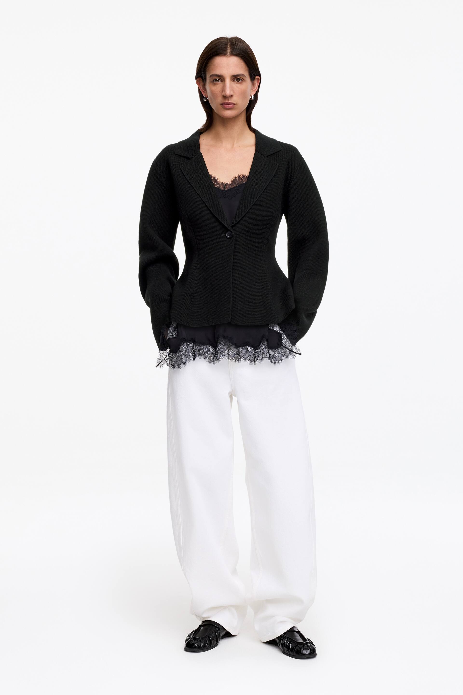 Visualizza immagine più grande: La giacca in maglia di lana-cotone nera è indossata dalla modella, caratterizzata da una texture a maglia visibile, revers a lancia, chiusura a un bottone in vita aderente e un sottile orlo a peplo. La giacca è sovrapposta a un top nero con bordi in pizzo.