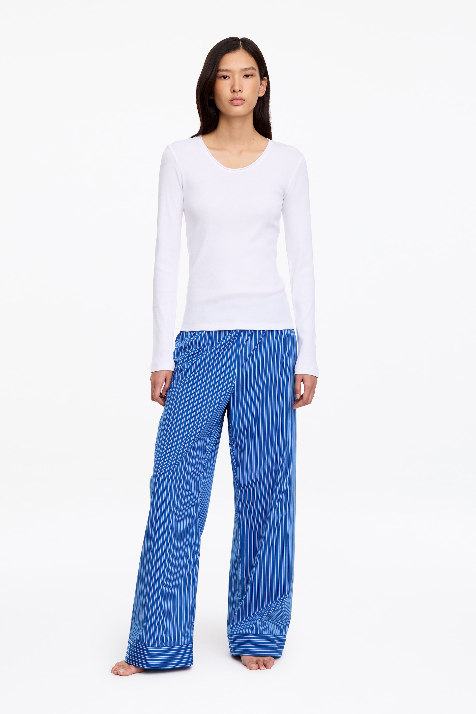 Größeres Bild anzeigen: Der Model trägt eine blau-weiß vertikal gestreifte Pyjamahose mit entspannter, weit geschnittener Silhouette. Die Hose verfügt über ein horizontal gestreiftes Detail am Saum und wird mit einem langärmeligen weißen Oberteil kombiniert.