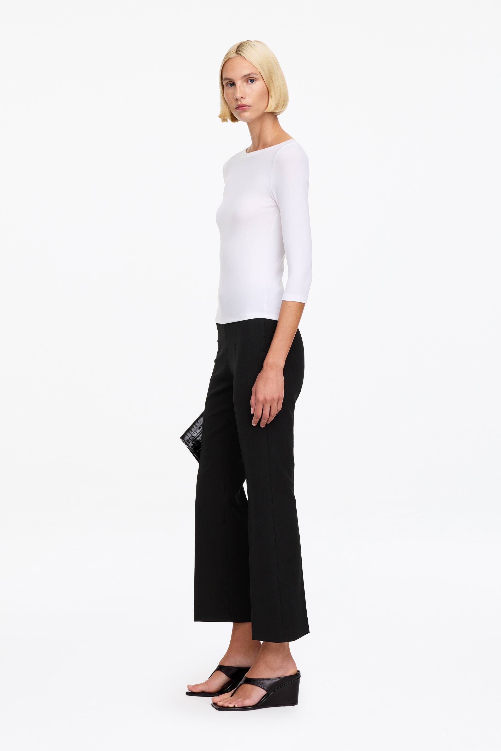 Ver imagen más grande: La modelo lleva un pantalón negro entallado, de corte slim y largo al tobillo. El pantalón se combina con un top blanco ajustado de manga tres cuartos, sandalias negras de cuña y un bolso de mano negro que lleva en una mano.