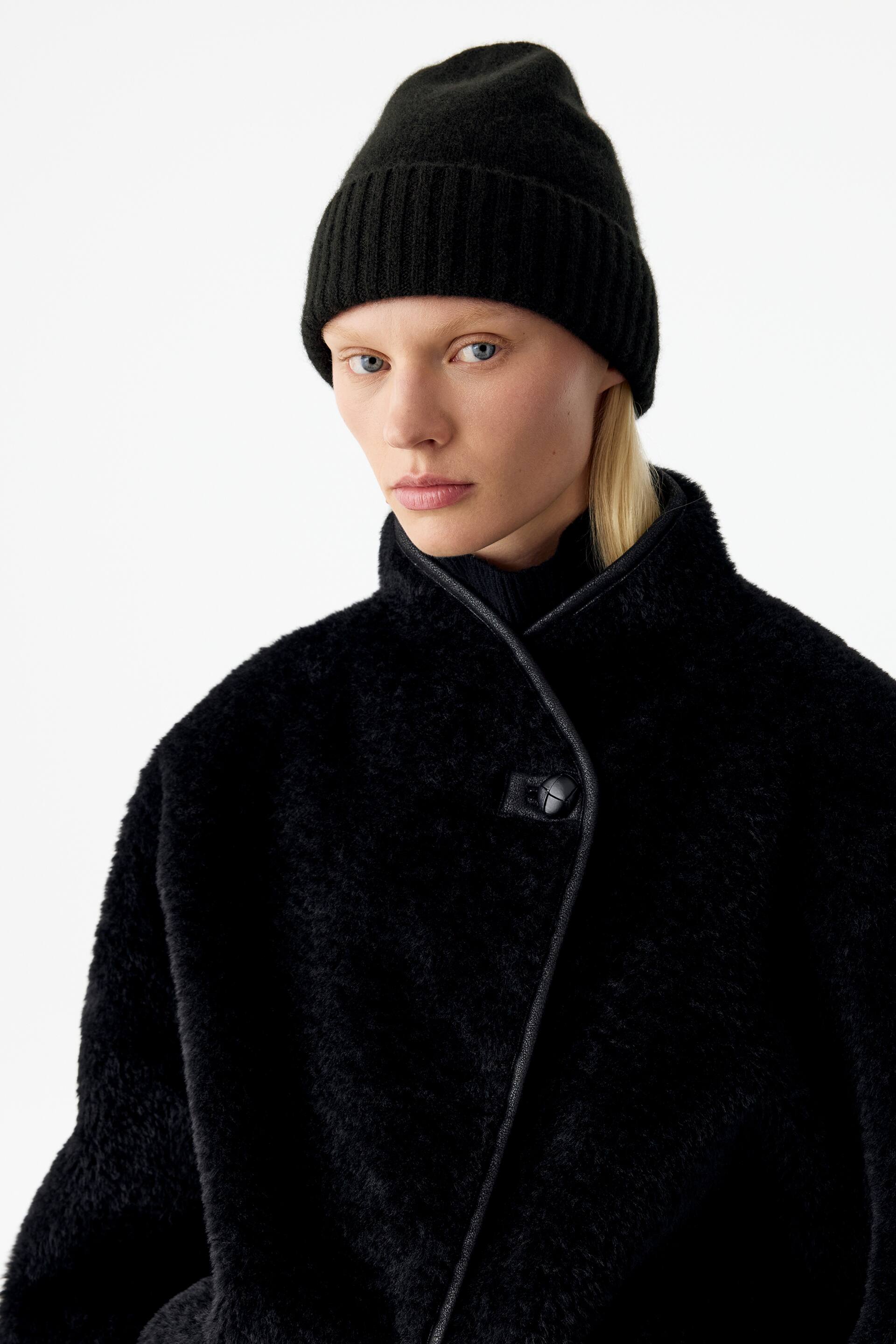 Grotere afbeelding bekijken: Een zwarte gebreide beanie met een brede, geribbelde omgeslagen rand wordt gedragen door het model. De aansluitende beanie wordt gecombineerd met een zwarte jas met textuur en hoge kraag.