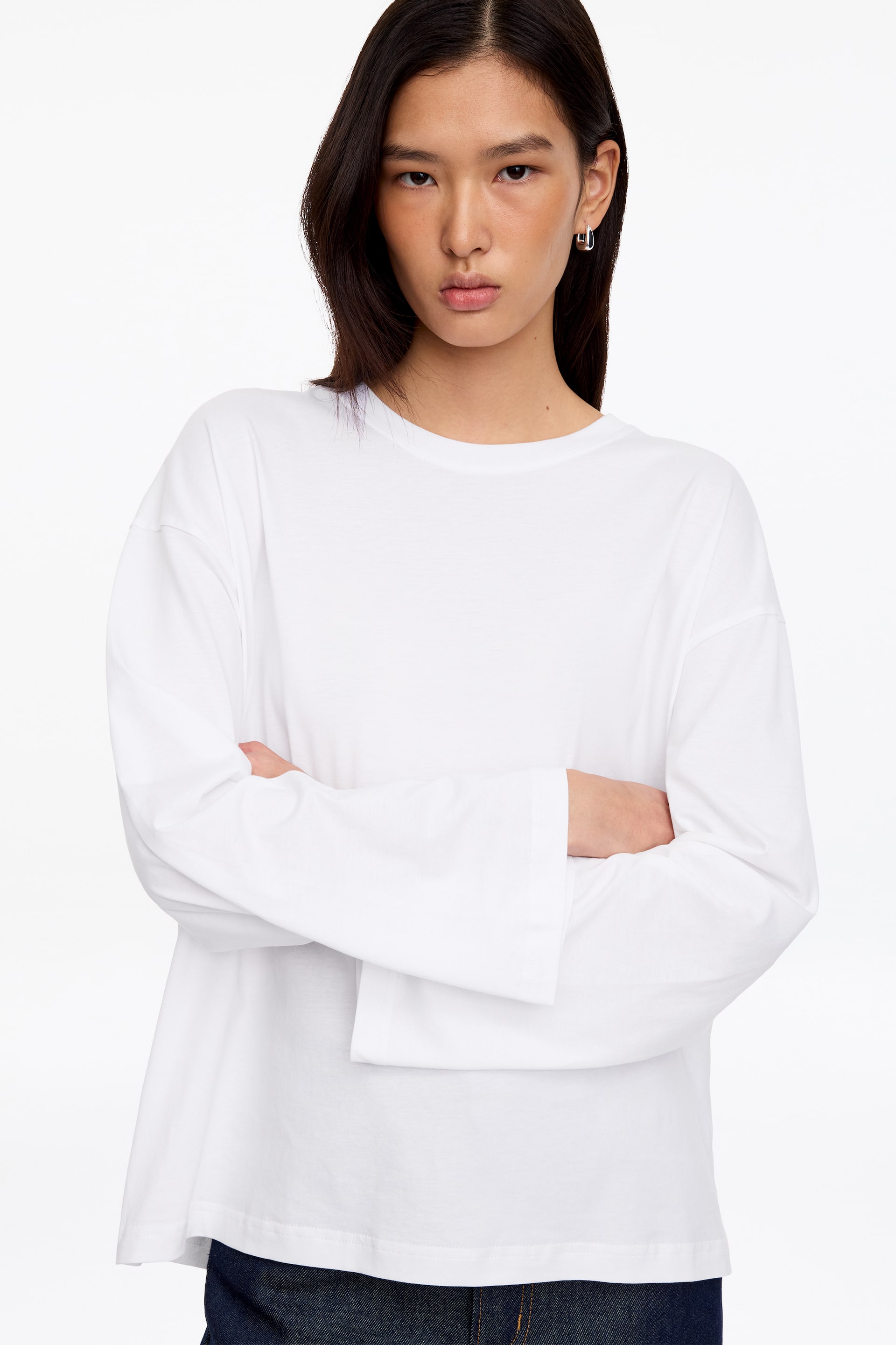 Visa större bild: En vit T-shirt med avslappnad passform, långa ärmar, rund halsringning och nedsänkta axlar bärs av modellen, som står med armarna i kors. T-shirten stylas med mörkblå denimbyxor som syns i midjan.