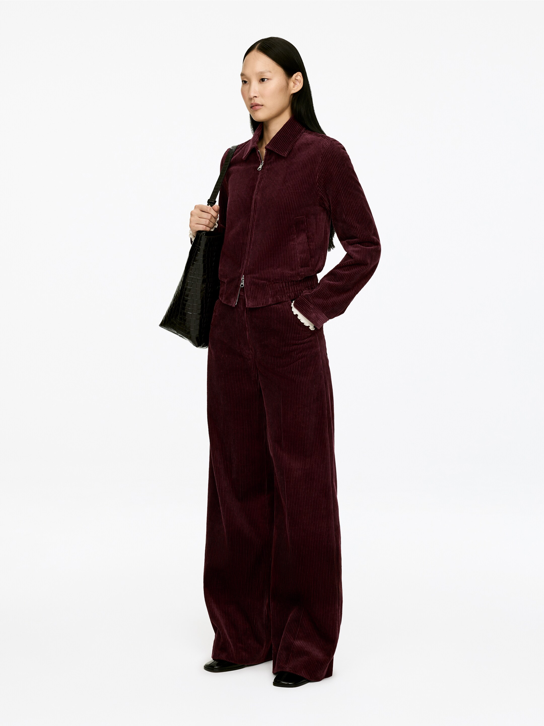 Ver imagen más grande: Un conjunto de dos piezas de pana burgundy se presenta desde un ligero ángulo frontal-derecho, con una chaqueta corta y pantalones de pierna ancha. La chaqueta incluye un cuello, un cierre frontal completo con cremallera, un dobladillo elástico y mangas largas con volantes blancos que asoman por los puños, mientras que los pantalones de pierna ancha a juego son de longitud completa.