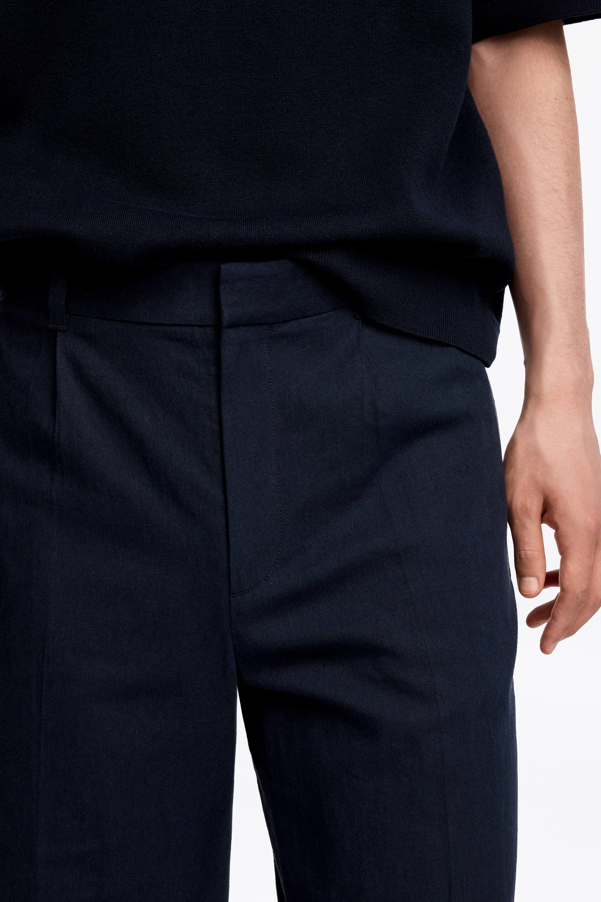 Grotere afbeelding bekijken: Donkerblauwe broek wordt afgebeeld vanuit een voor-rechts hoek, met een vlakke voorkant met zichtbare tailleband, riemlussen en een enkele plooi. Een donkerblauwe gebreide top met korte mouwen wordt over de tailleband van de broek gedragen.