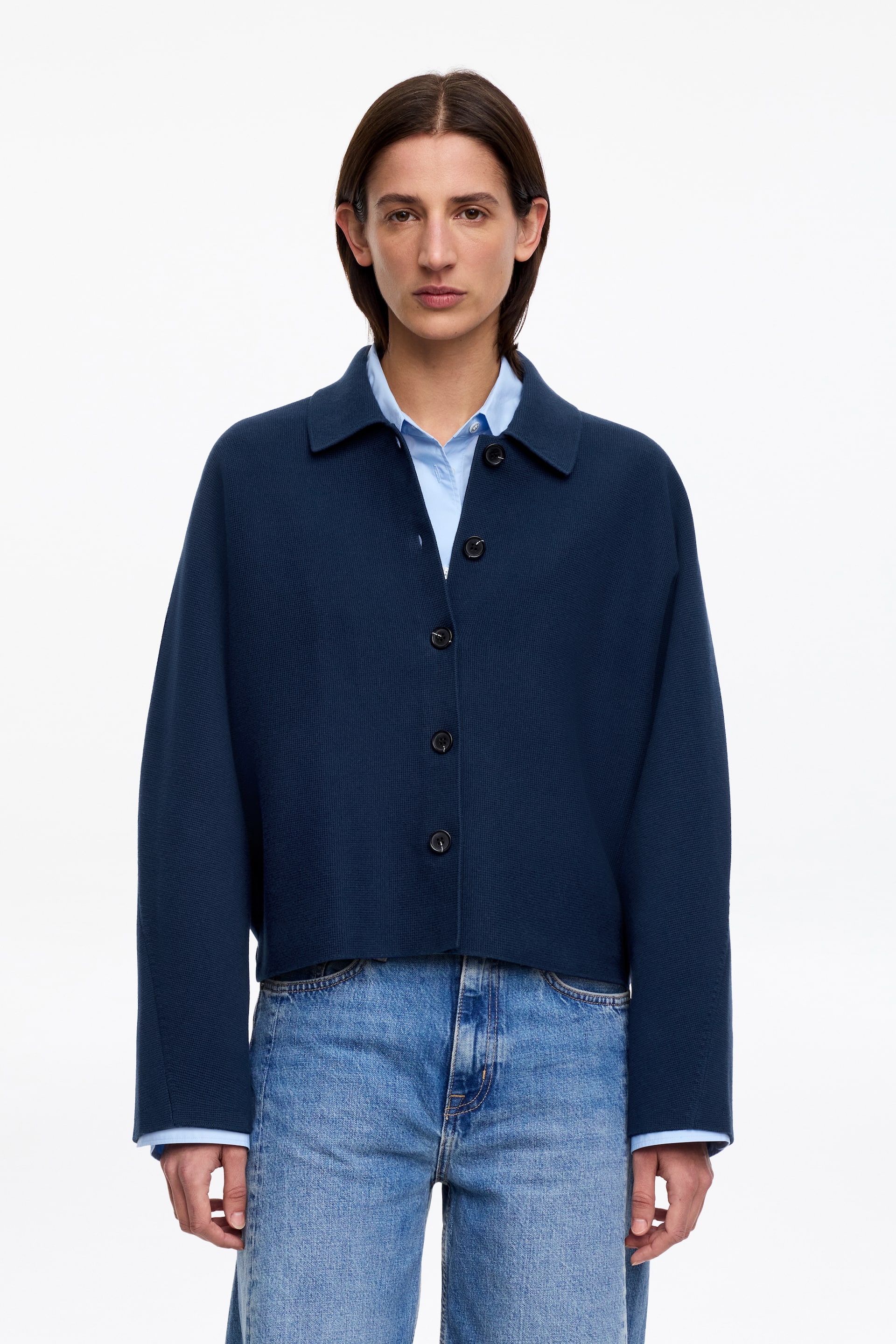 Agrandir l'image: Une veste en tricot bleu marine raccourcie avec un col pointu et une fermeture frontale à cinq boutons. Elle est portée par-dessus une chemise à col bleu clair, avec le col et les poignets de la chemise visiblement dépassant.
