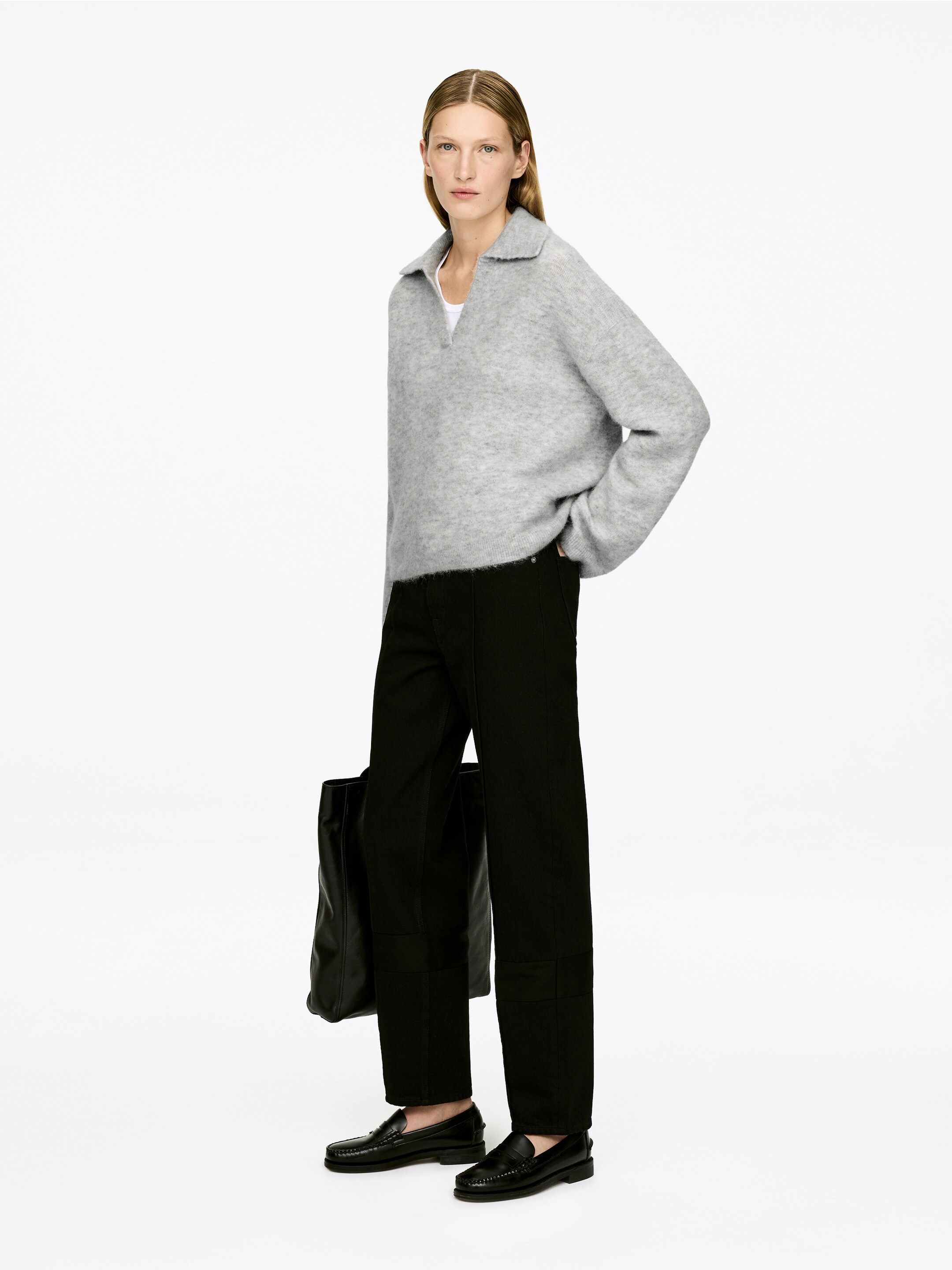 Größeres Bild anzeigen: Ein grauer Strickpolo-Pullover wird aus einem leichten Front-Links-Winkel präsentiert. Er verfügt über eine entspannte Passform mit einem spitzen Kragen und einem V-Ausschnitt, unter dem ein weißes T-Shirt sichtbar ist.