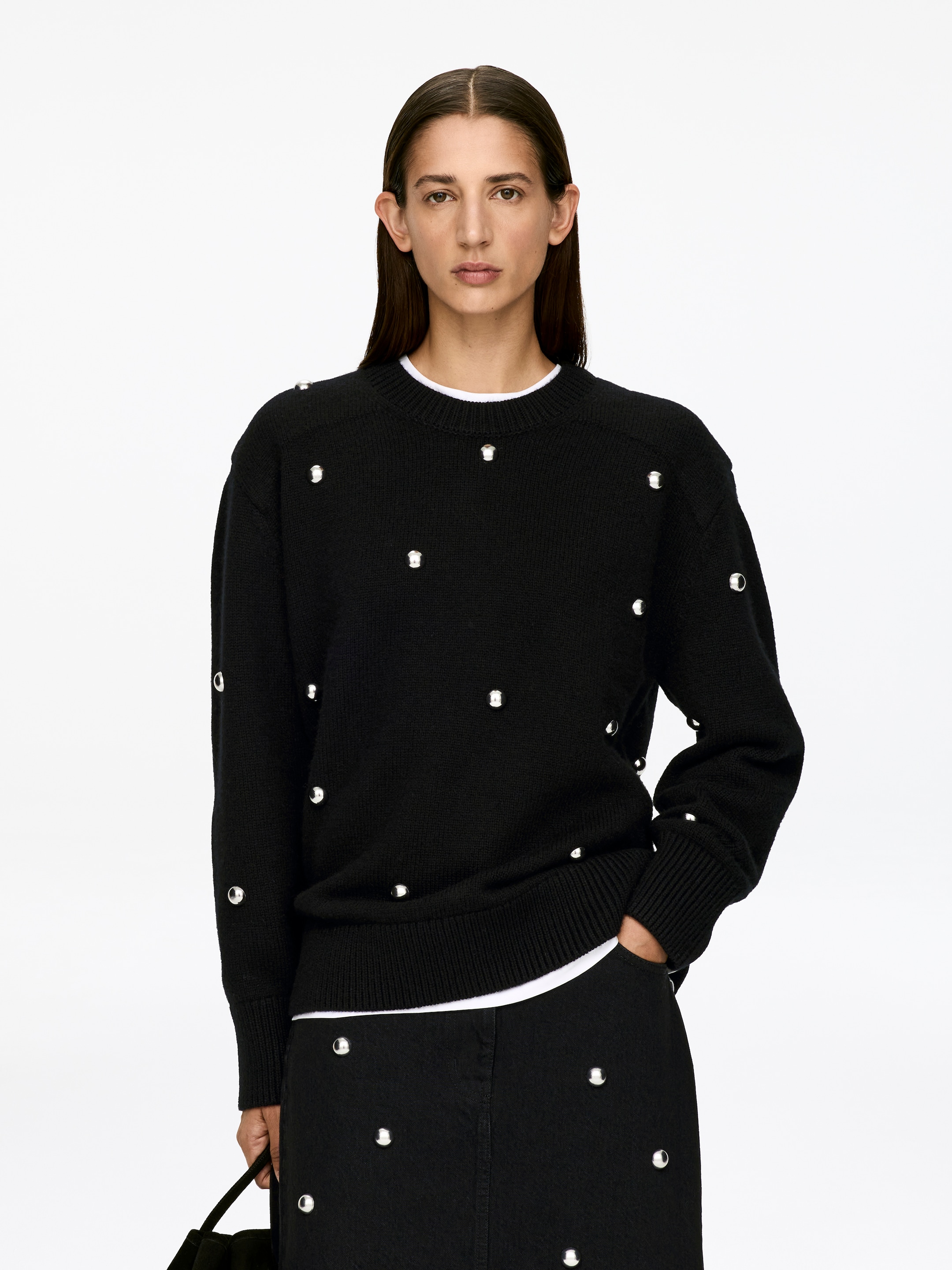 Grotere afbeelding bekijken: Een zwarte trui met ronde hals, voorzien van geribbelde boordafwerkingen en verspreide zilverkleurige studs, wordt door het model gedragen over een wit t-shirt, dat zichtbaar is bij de halslijn en zoom. De hand van het model rust in de zak van zwarte broek, die ook voorzien zijn van studs detailing.