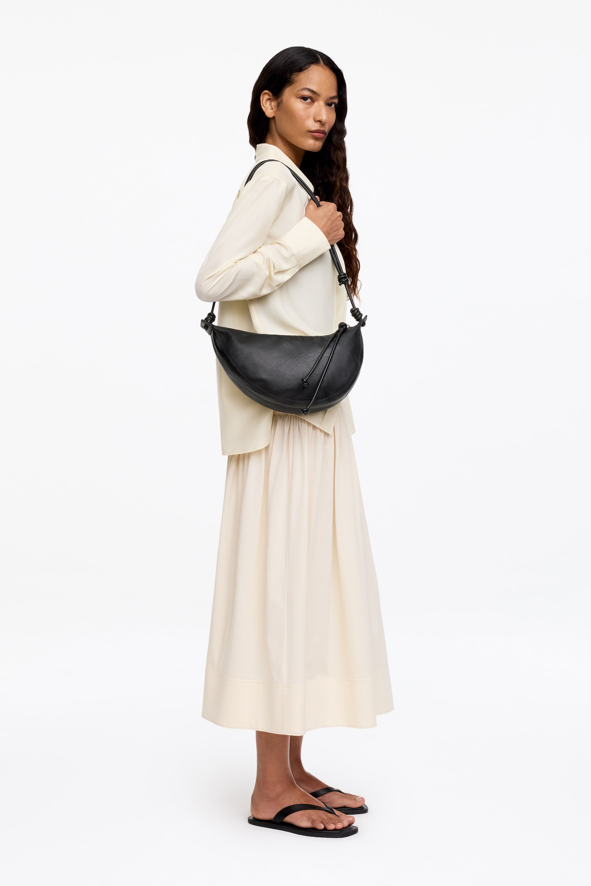 Ver imagem maior: Uma mala preta em forma de crescente é usada atravessada no corpo, repousando contra a anca, apresentando uma silhueta suave e curva, uma alça fina com anexos em nó e um detalhe decorativo em laço na sua superfície frontal.