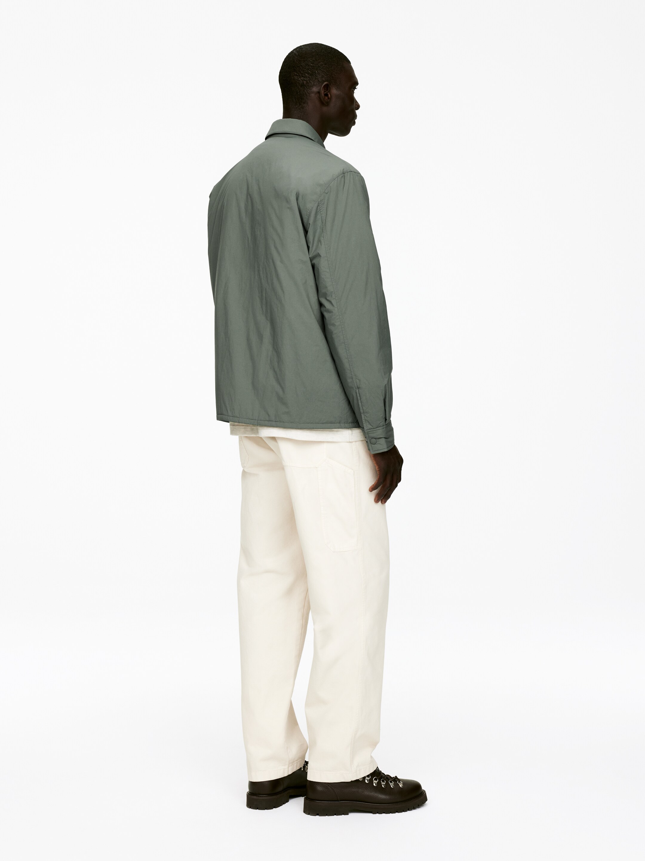 Ver imagen más grande: Una chaqueta ligera de color verde apagado con un cuello en punta y un ajuste relajado se muestra desde una vista trasera, superpuesta sobre una camiseta blanca. Unos pantalones de pierna ancha de color crema con costuras laterales visibles y bolsillos de parche se llevan puestos, combinados con botas marrón oscuro con suelas gruesas y ojales de anillo en D plateados.