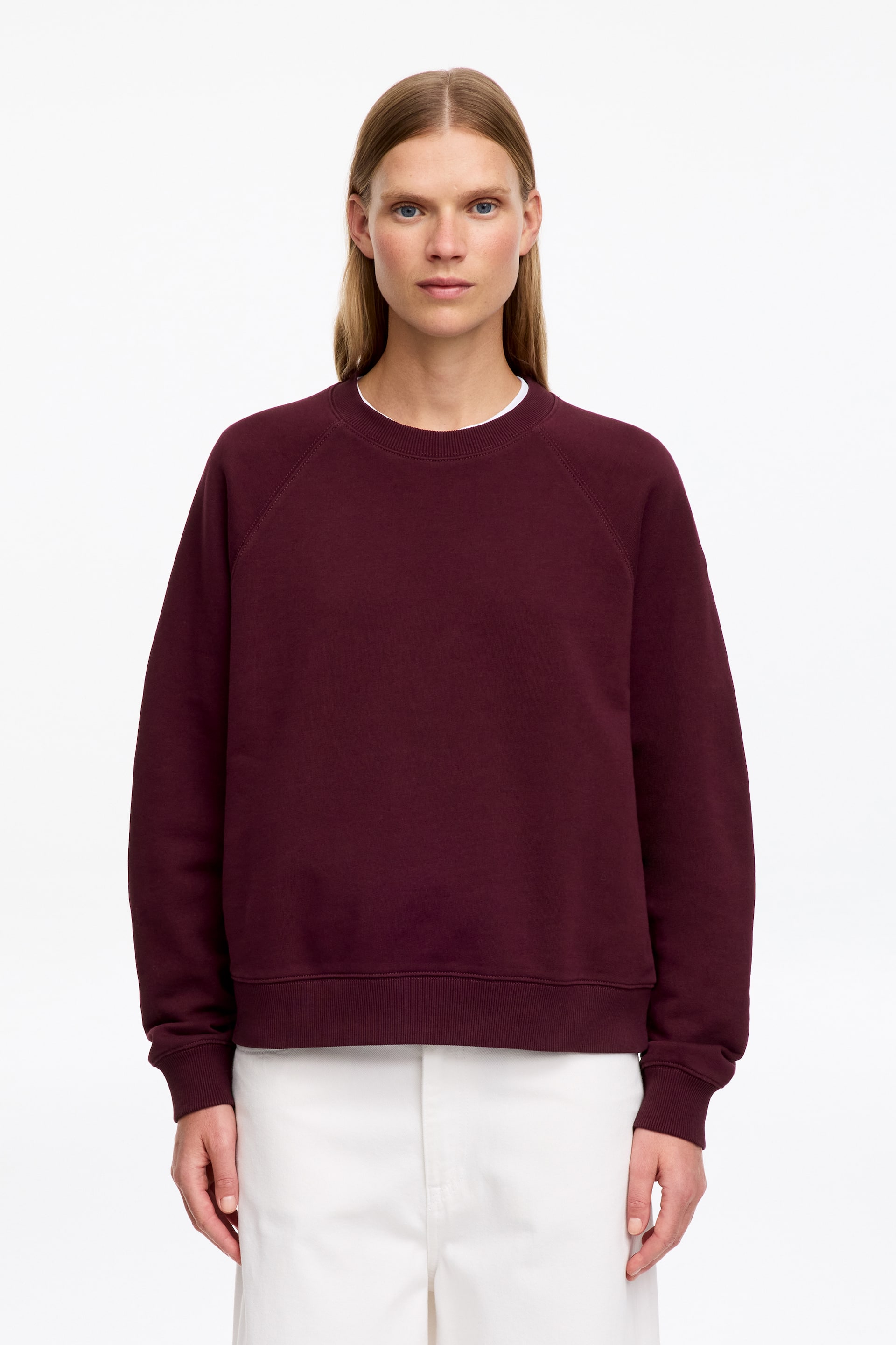 Agrandir l'image: Un sweat-shirt col rond bordeaux est porté par le mannequin, avec des manches raglan et des finitions côtelées au col, aux poignets et à l’ourlet. Le sweat-shirt est porté sur un t-shirt blanc et associé à un pantalon blanc.