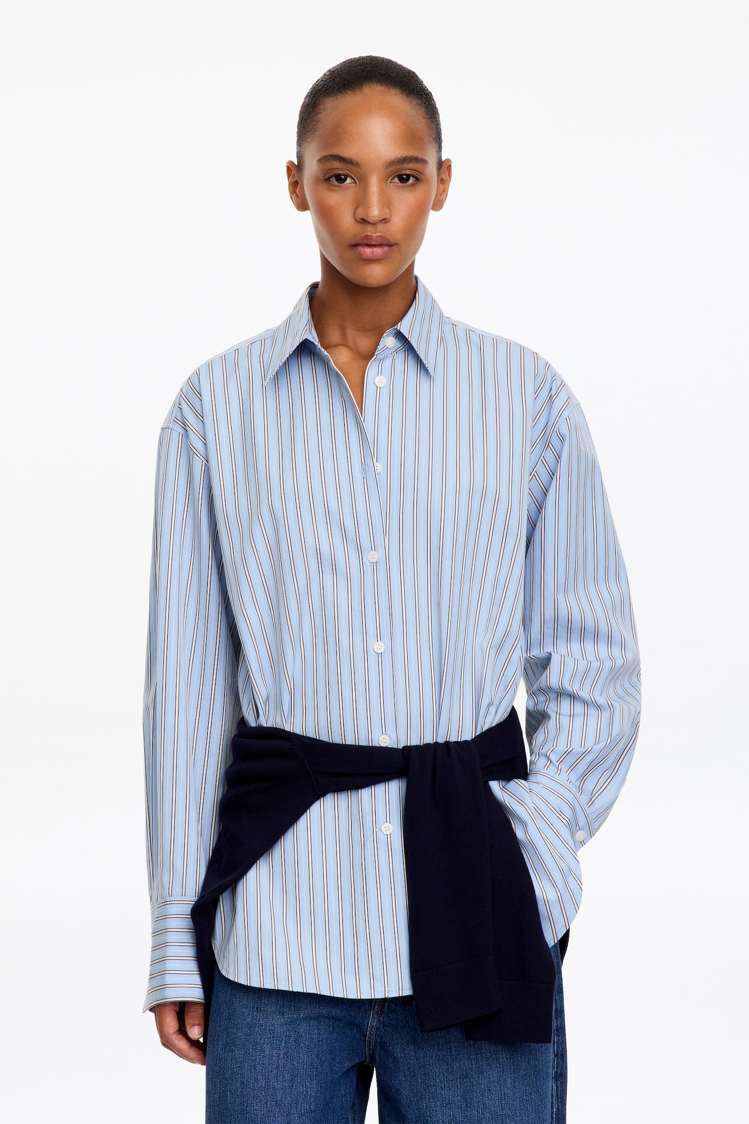 Una camisa de manga larga y abotonada presenta desde el frente un estampado de finas rayas verticales en azul claro. La camisa luce una silueta relajada con hombros caÃdos, un clásico cuello de punta, puños anchos y un bajo curvado. - 1