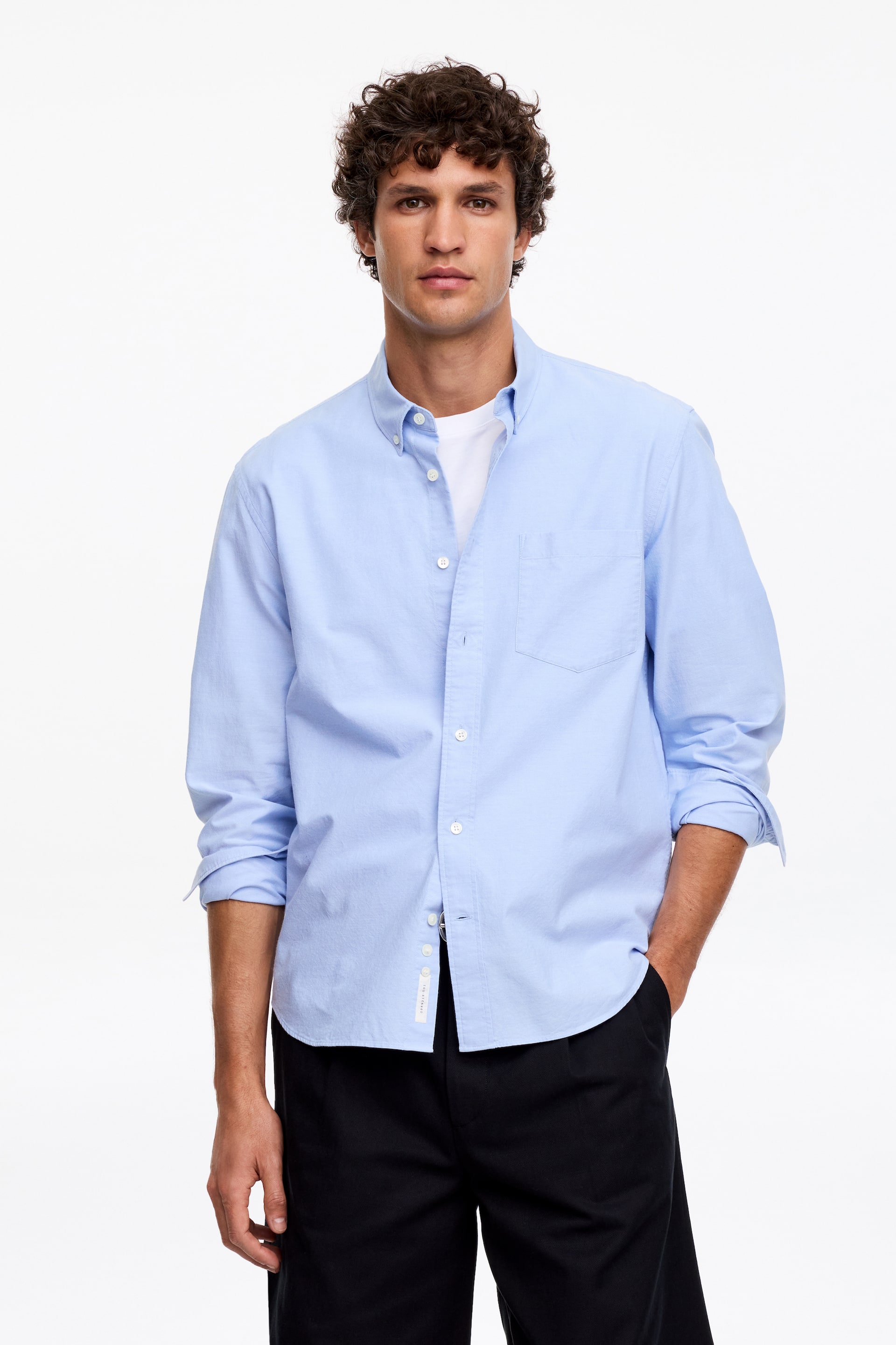 Zobacz większy obraz: Jasnoniebieska koszula z długim rękawem, zapinana na guziki, jest widoczna z przodu. Posiada kołnierzyk zapinany na guziki, białe guziki biegnące wzdłuż przodu oraz pojedynczą kieszeń na piersi po lewej stronie. Rękawy są podwinięte do przedramion, a pod spodem widoczny jest biały t-shirt.