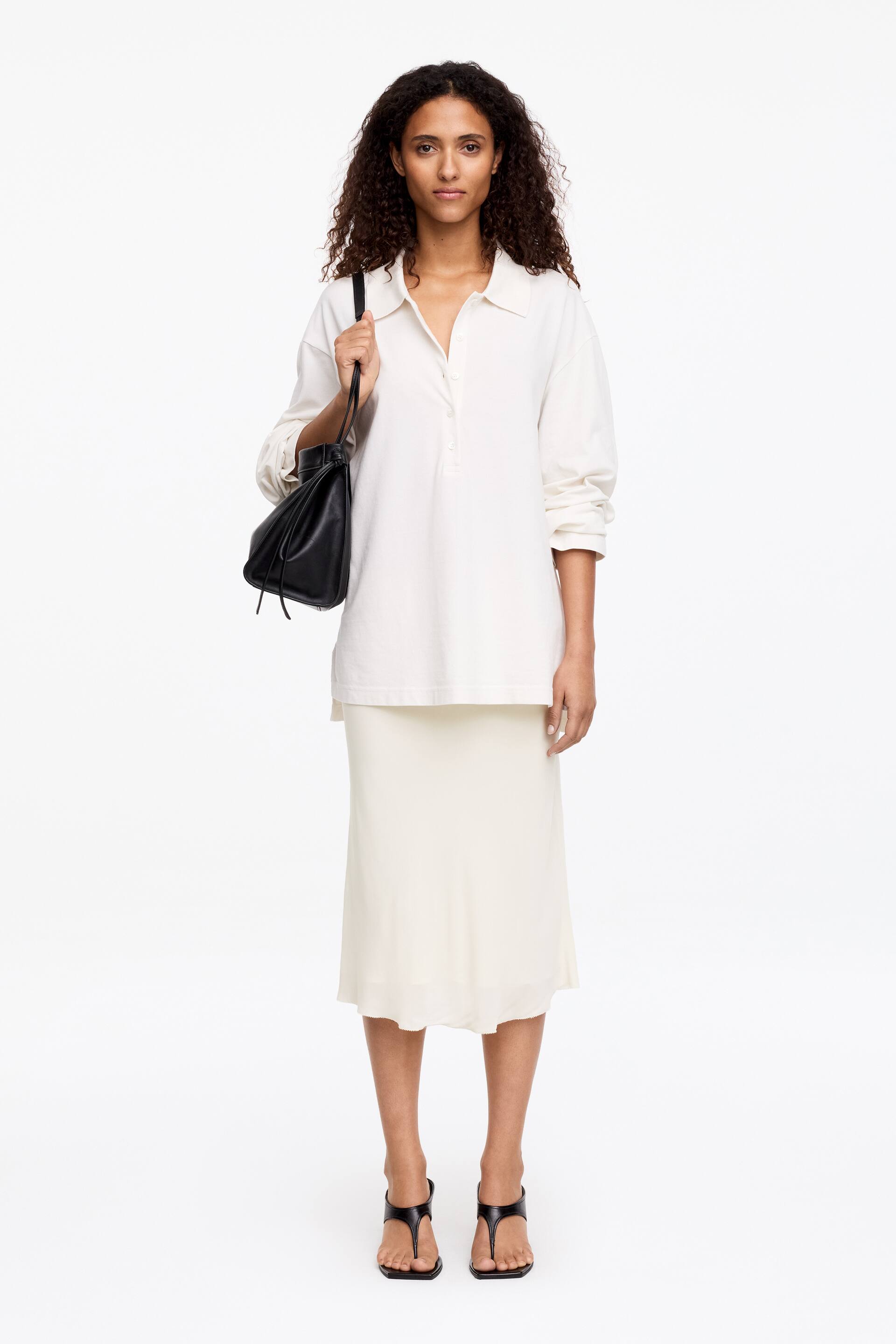 Grotere afbeelding bekijken: Een crèmekleurige midi-rok van dubbele laag chiffon wordt gedragen door het model, vallend in een zachte, vloeiende vorm en gestyled onder een crèmekleurige poloshirt met lange mouwen.