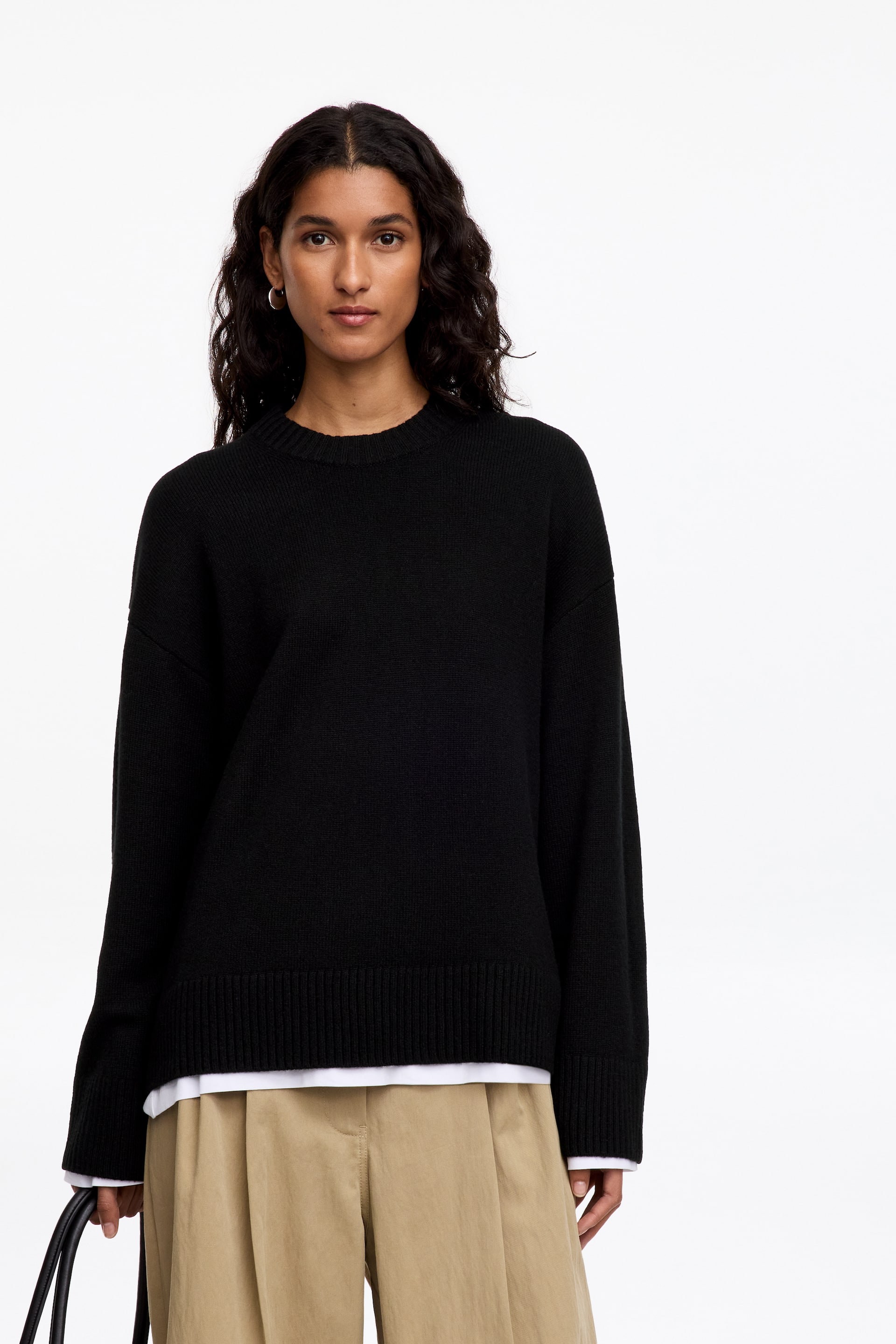 Visualizza l'immagine ingrandita: Una maglia nera a girocollo in maglia, caratterizzata da spalle scese e bordi a coste, indossata dalla modella. La maglia dalla vestibilità rilassata è abbinata sopra una camicia bianca e completata con pantaloni beige.
