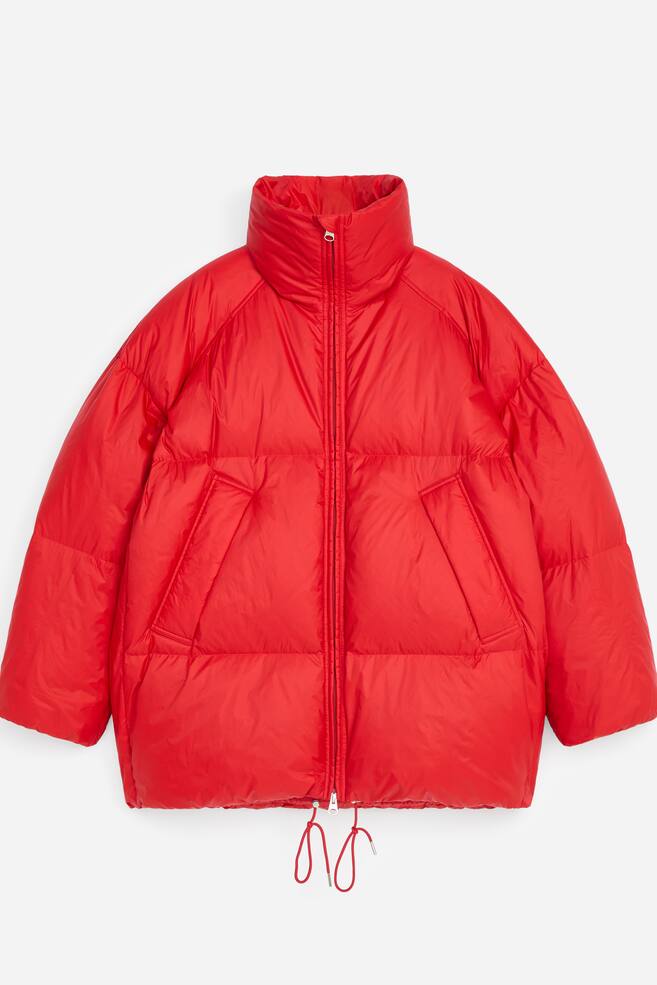Eine leuchtend rote Steppjacke ist von vorne zu sehen und zeigt eine voluminÃ¶se, oversized Silhouette mit horizontaler Steppung und einem hohen Stehkragen. Die Jacke verfÃ¼gt Ã¼ber einen durchgehenden FrontreiÃverschluss, zwei groÃe schrÃ¤ge Paspeltaschen und rote Kordeln, die am unteren Saum herunterhÃ¤ngen. - 1