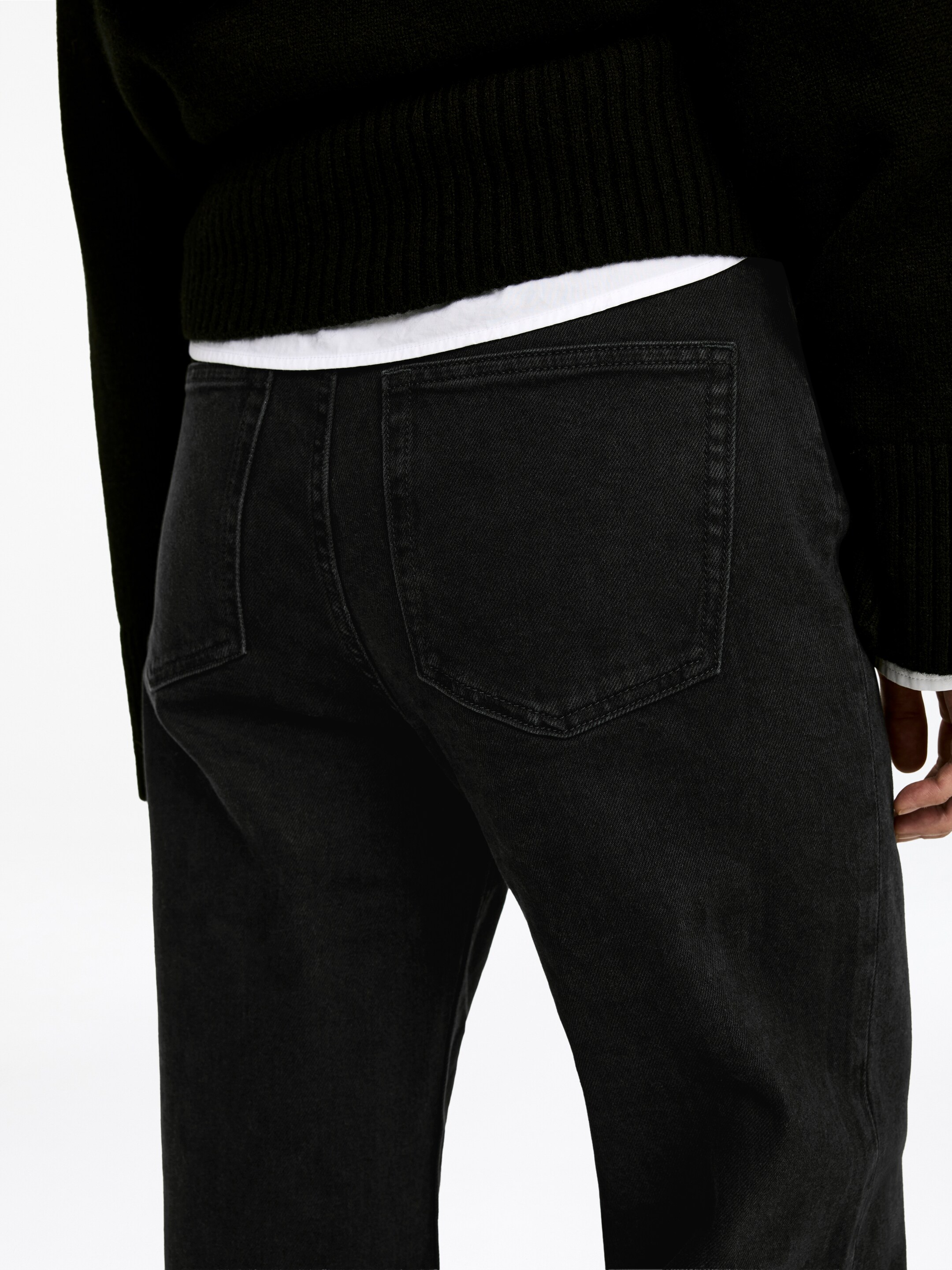 Größeres Bild ansehen: Schwarze Jeans werden aus einer hinteren, leicht schrägen Ansicht präsentiert, mit zwei rechteckigen Aufgesetzttaschen mit sichtbaren Nähten und einem verblichenen dunklen Waschton. Ein schwarzer gerippter Strickpullover deckt teilweise den Bund ab, darunter schichtet sich ein weißes T-Shirt.