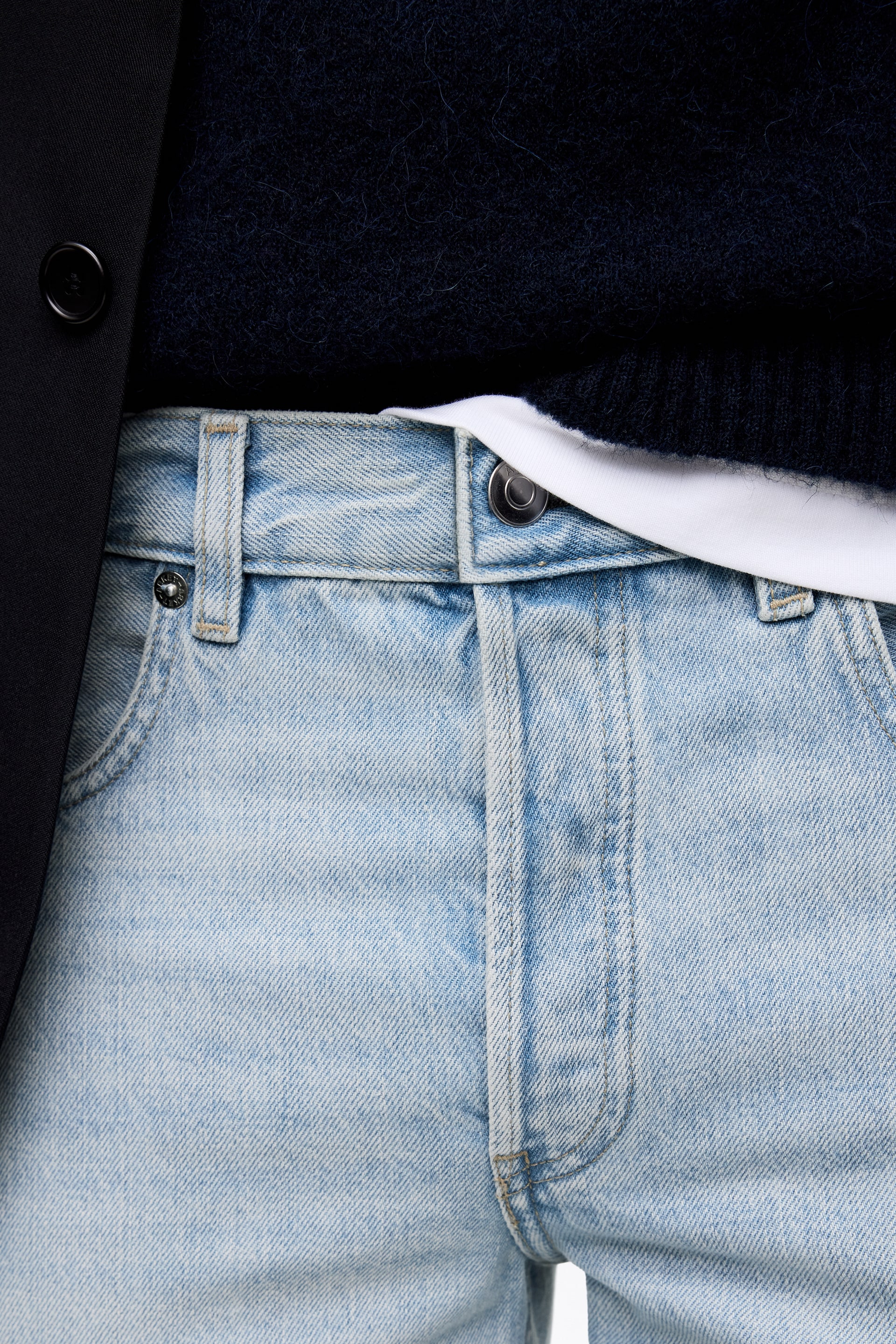Ver imagen más grande: Los vaqueros de mezclilla de lavado claro se muestran desde un ángulo frontal, con un cierre de botón metálico en la cintura, trabillas visibles y remaches metálicos cerca de los bolsillos delanteros. Un suéter azul oscuro se coloca sobre una camiseta blanca, ambos metidos en los vaqueros, con una chaqueta negra cubriendo parcialmente el lado izquierdo.