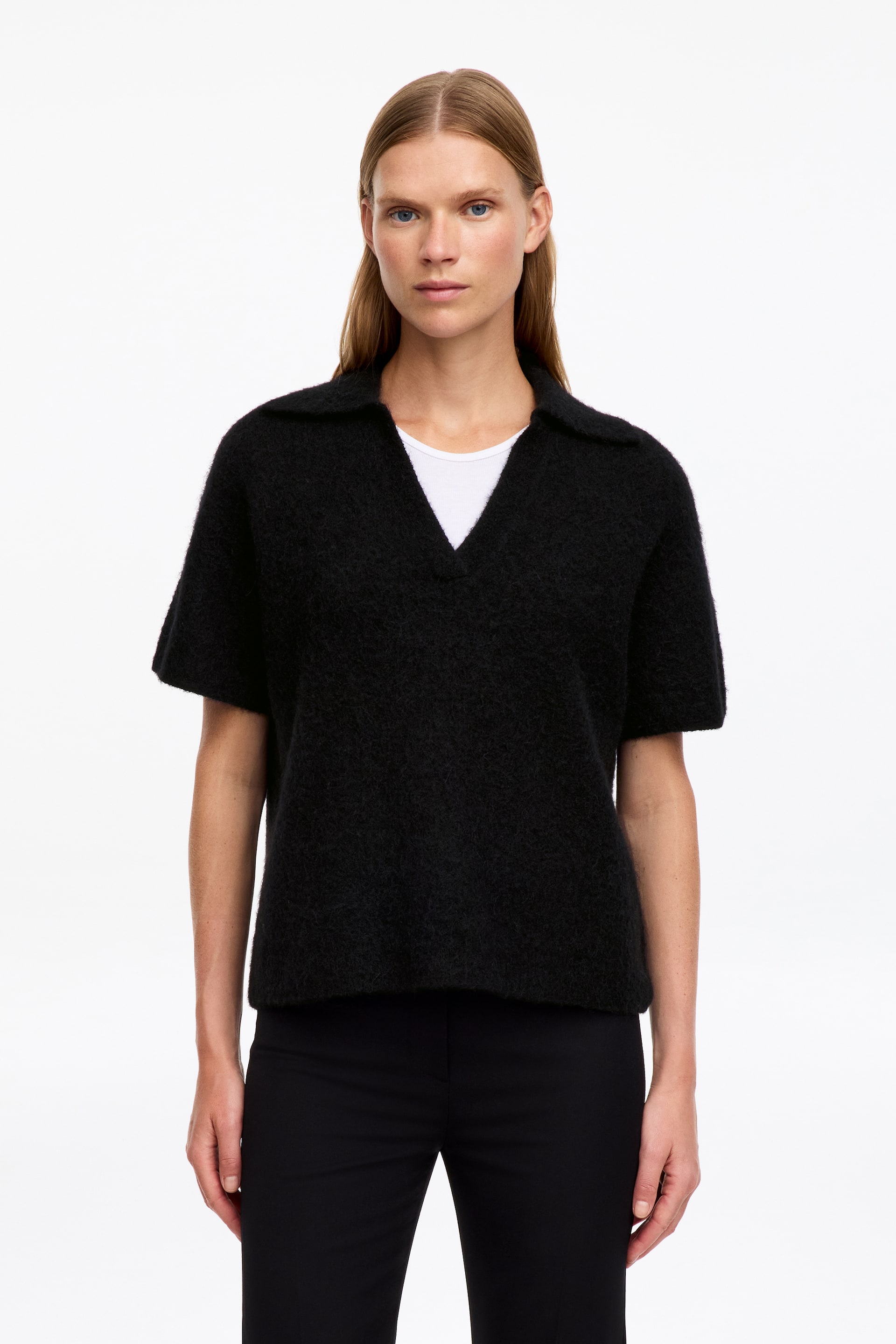 View larger image: A black polo jumper, featuring short sleeves, an open V-neck and a textured knit, is being worn by the model. The jumper has a loose fit, with a white T-shirt visible underneath, while black trousers are also being worn.