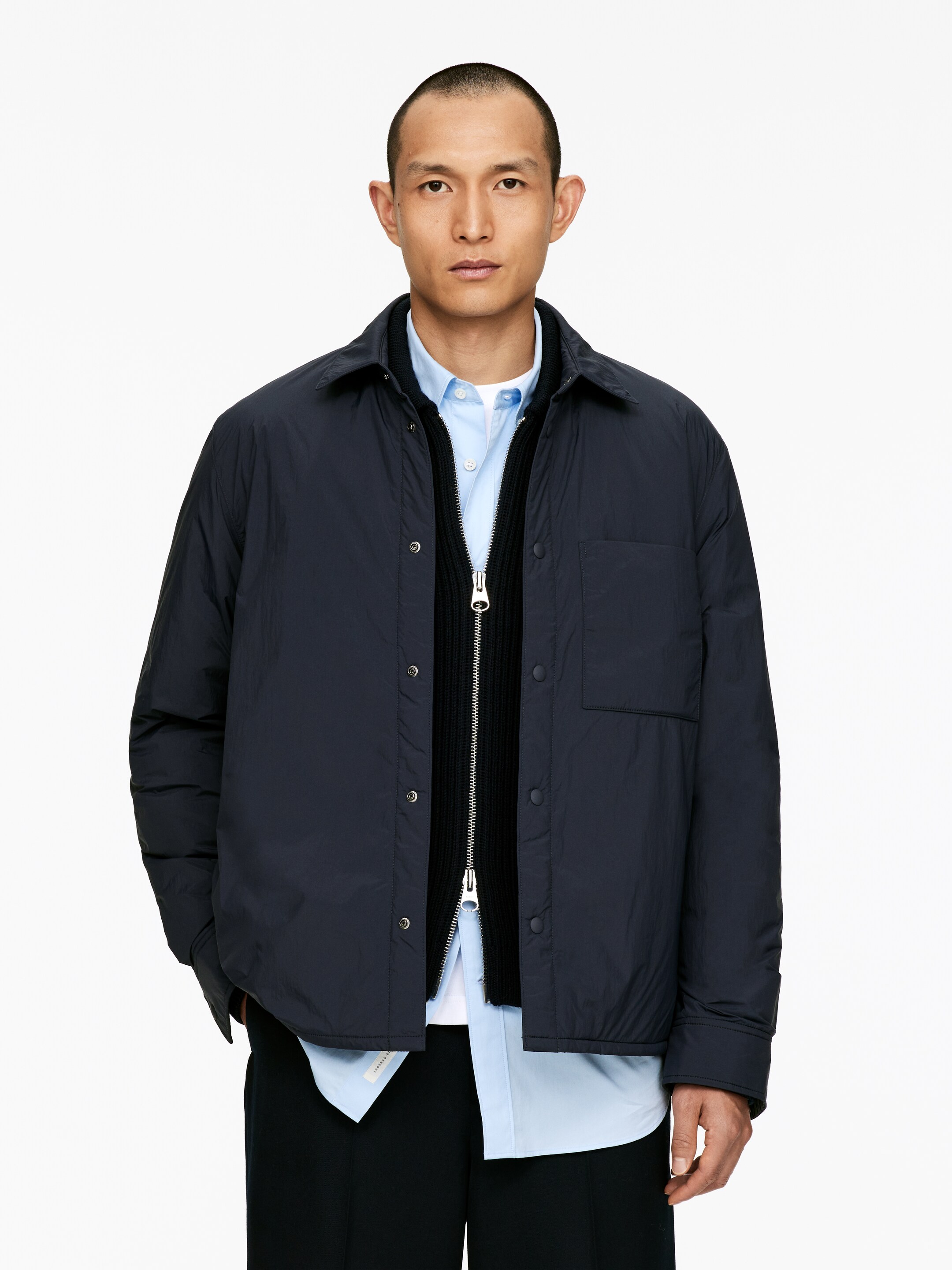 Visualizza immagine più grande: Una giacca camicia imbottita blu navy è mostrata su un modello rivolto in avanti. La giacca presenta un colletto classico, chiusure a bottoni a pressione lungo la parte anteriore e una singola tasca a toppa sul petto destro di chi la indossa, ed è indossata aperta sopra un maglione nero con zip e una camicia azzurra con colletto.