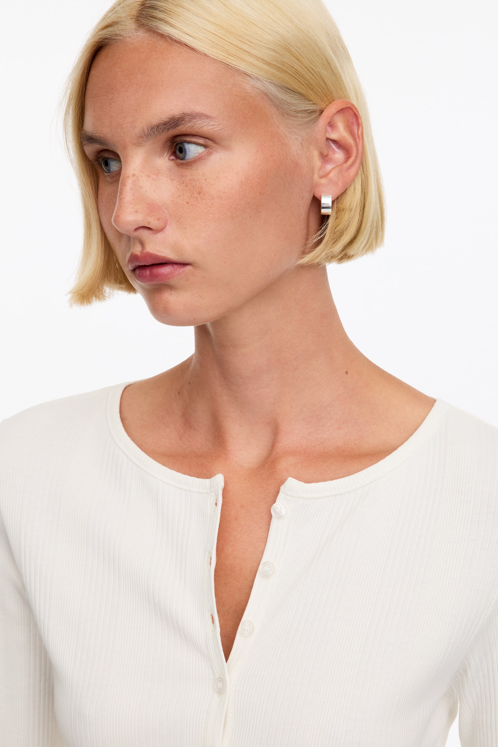 Visualizza immagine più grande: Un top color crema in maglia a coste è indossato dalla modella, caratterizzato da uno scollo tondo e da una stretta abbottonatura frontale con piccoli bottoni delicati lasciata parzialmente aperta.