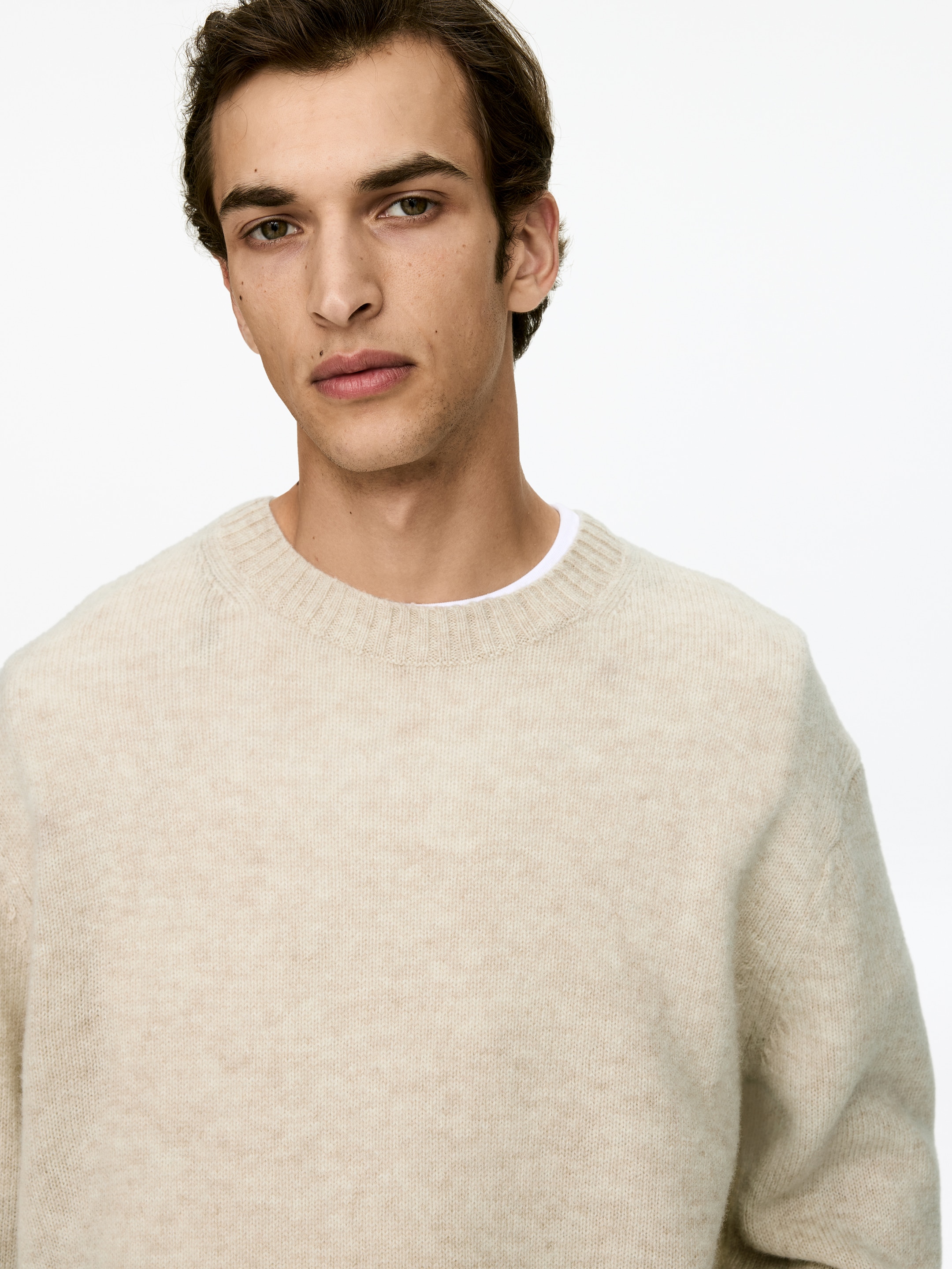 Visa större bild: En ljusbeige tröja med rund hals visas från en framåtvänd vinkel, med en mjuk, subtilt texturerad stickning och en ribbad krage. En vit t-shirt syns som ett lager under tröjans halsringning.