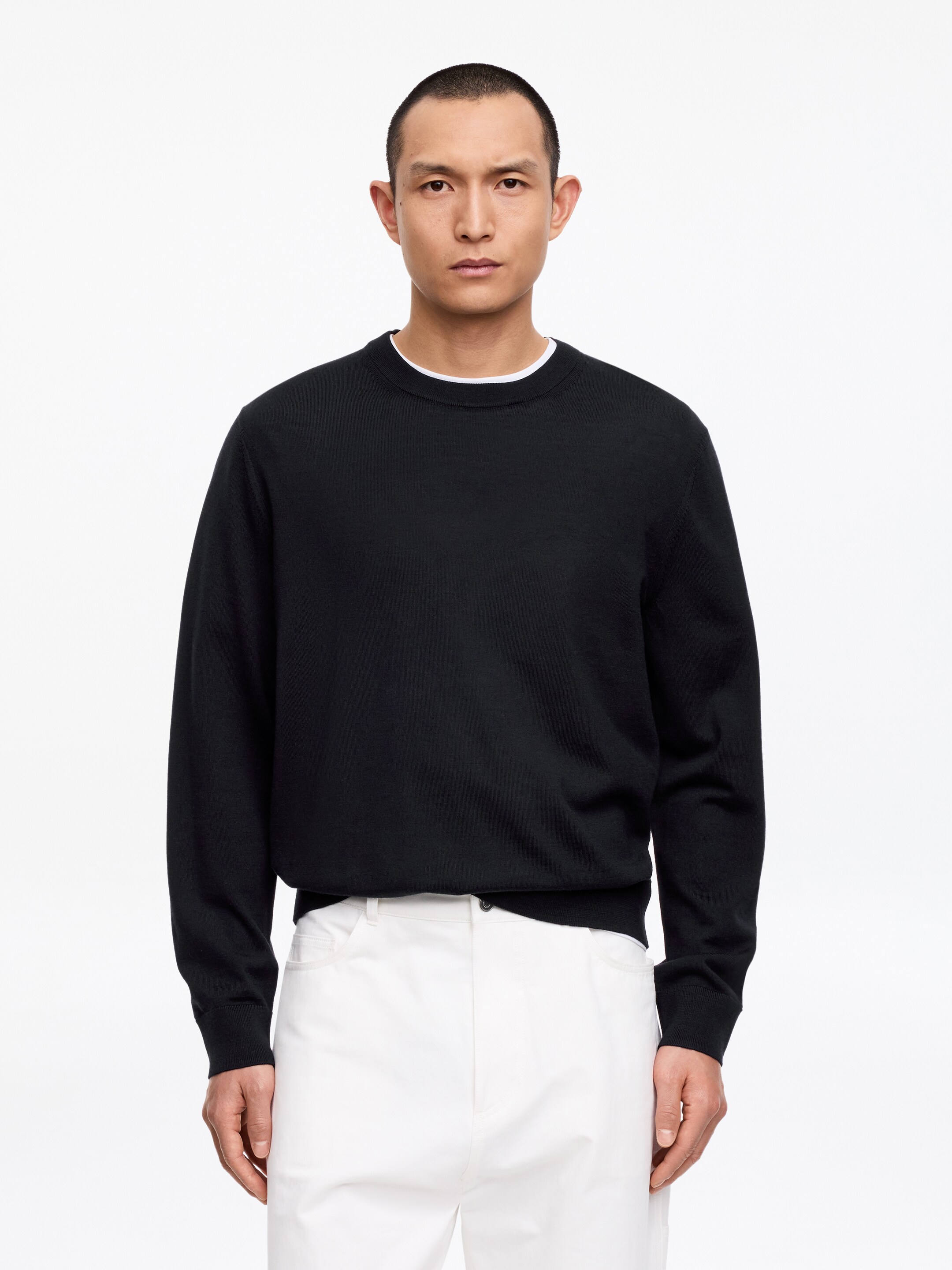 Grotere afbeelding bekijken: Een zwarte sweater met ronde hals wordt gepresenteerd, met lange mouwen, geribde manchetten en een geribde boord, waarbij een witte T-shirtkraag zichtbaar is onder de halslijn.