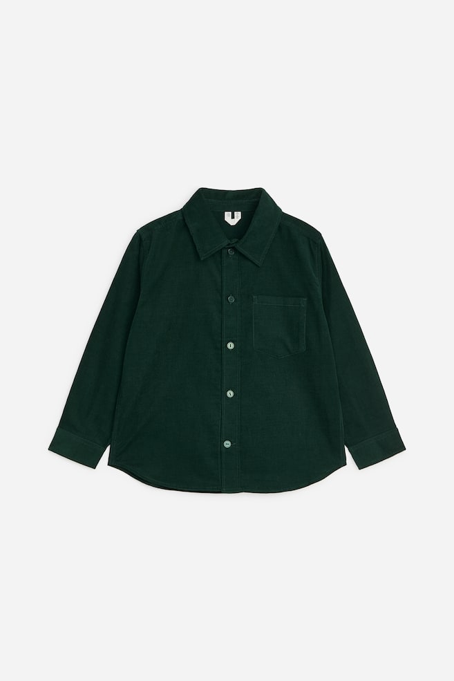 Una camicia in velluto a coste di cotone a maniche lunghe, di colore verde scuro, è appoggiata in piano in una vista frontale, mostrando un colletto a punta, una chiusura frontale con bottoni, una singola tasca sul petto, polsini abbottonati e un orlo arrotondato. - 1