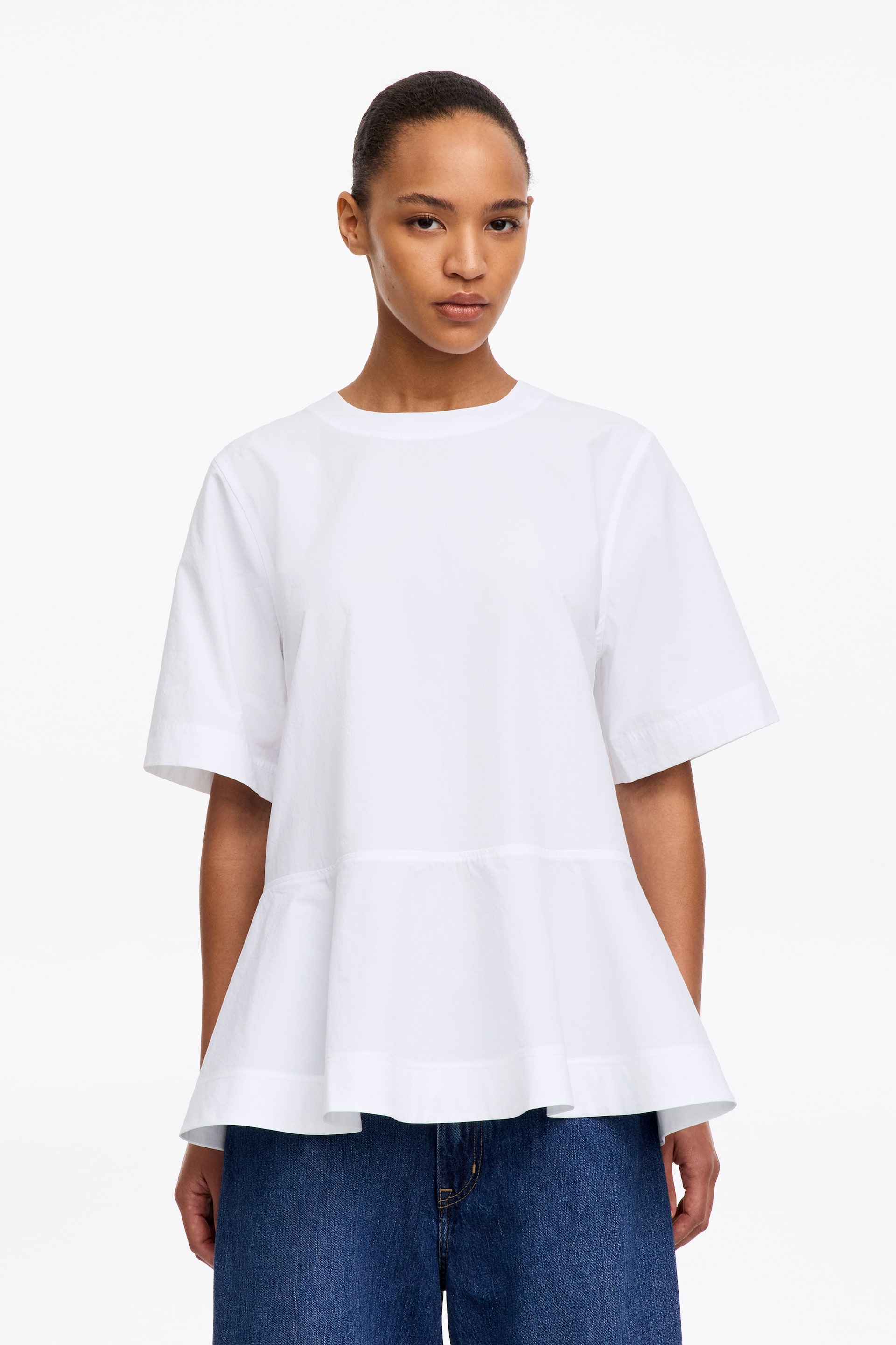 Ver imagem maior: A modelo está a usar uma T-shirt Peplum em Algodão Pima branco. Apresenta gola redonda, mangas curtas e uma bainha peplum evasé, combinada com jeans de ganga azul.