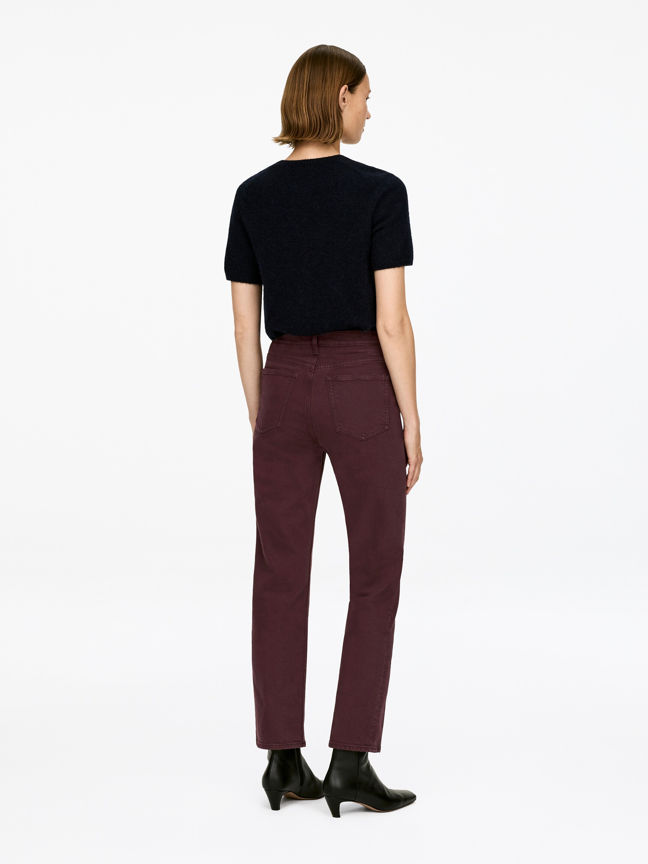 Vezi imaginea mai mare: Pantalonii JADE CROPPED de un roșu închis sunt purtați de model, având talie medie, o croială slim și picioare înguste și crop. Pantalonii cad chiar deasupra unor cizme negre de gleznă, subliniind lungimea lor.